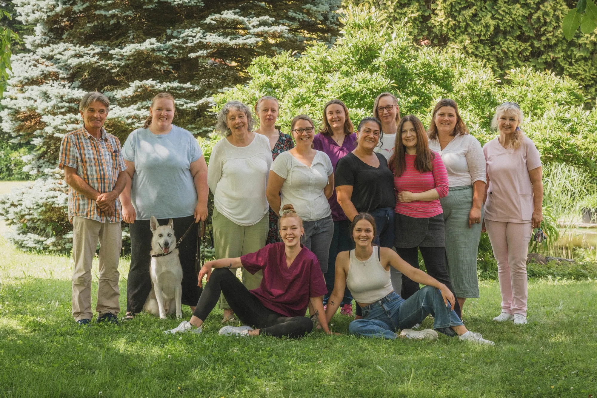 Gruppenfoto von zwölf Personen und einem weißen Hund auf einer grünen Wiese vor Büschen und Bäumen.