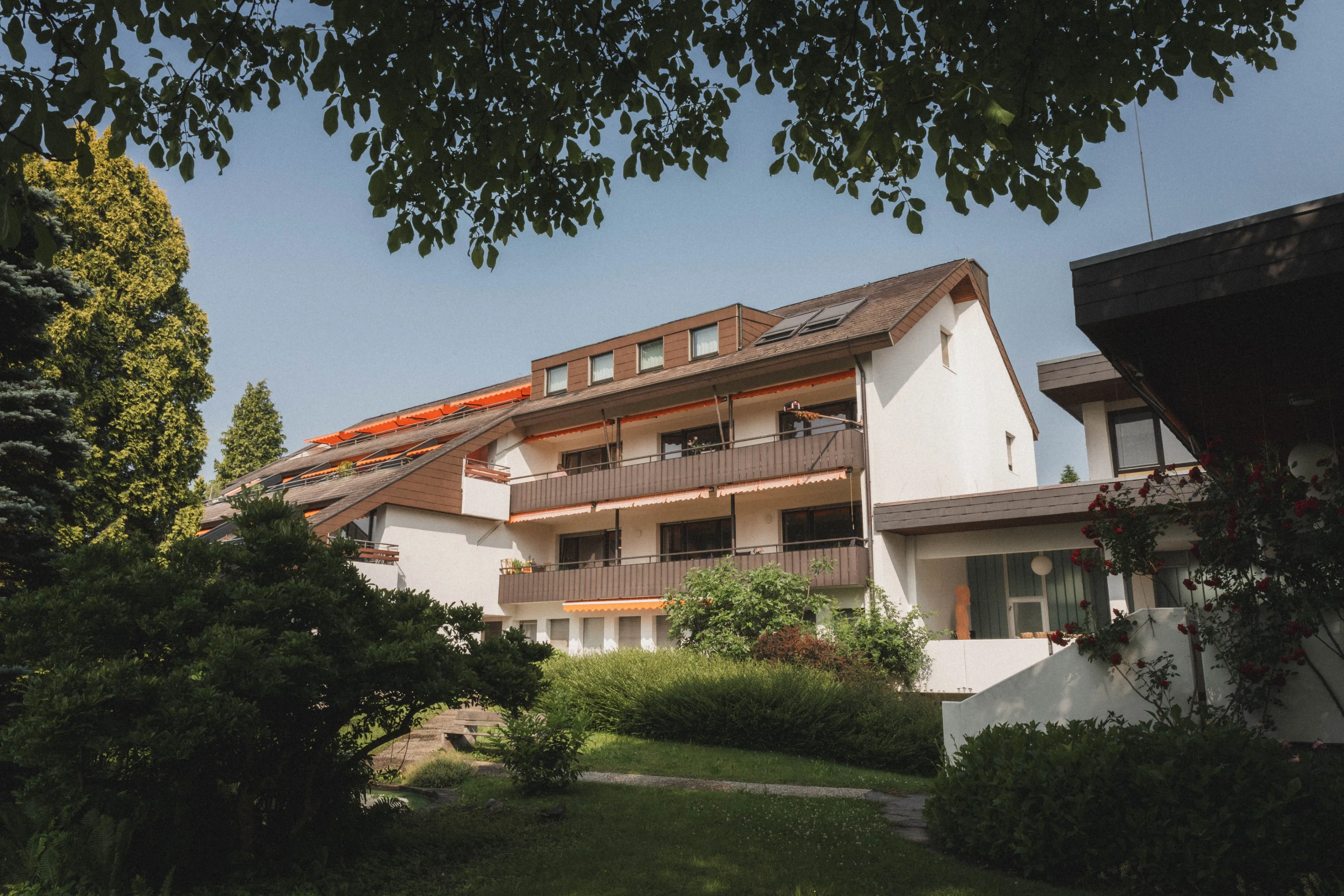 Mehrstöckiges Wohngebäude mit Balkonen und orangen Markisen, umgeben von grünen Bäumen und Sträuchern unter blauem Himmel.