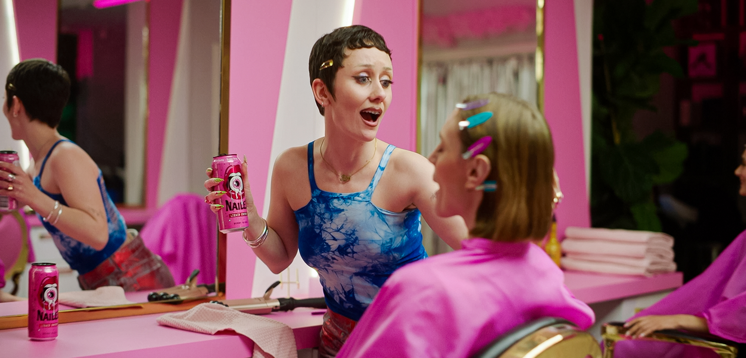 Woman in blue tie-dye top holding a pink NAILED energy drink can, talking to a seated woman with hair clips in a salon with pink decor.