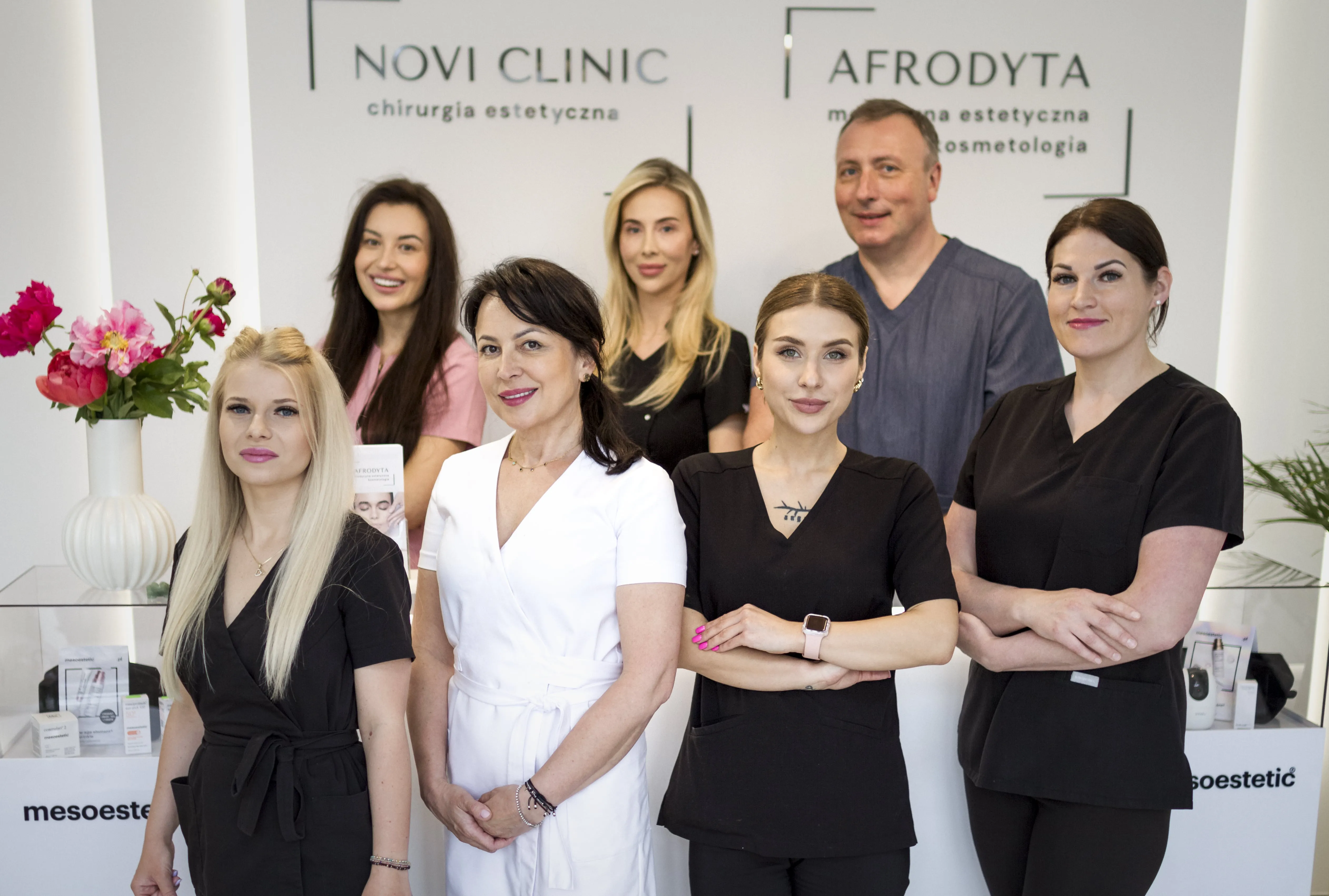 Zespół specjalistów kliniki medycyny estetycznej pozuje do zdjęcia w nowoczesnym wnętrzu gabinetu - uśmiechnięte kobiety i mężczyzna stoją przed logotypami "Novi Clinic" i "Afrodyta".
