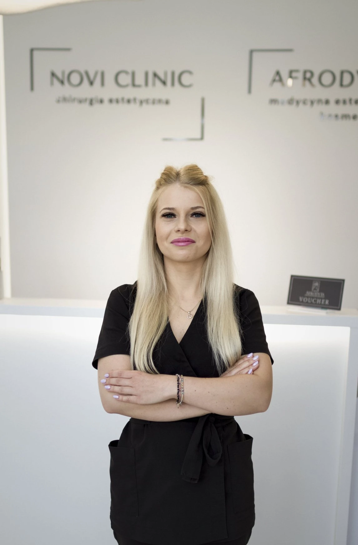 Kobieta w czarnym uniformie medycznym z długimi blond włosami stoi z założonymi rękami na tle recepcji kliniki. W tle widoczne logotypy „Novi Clinic” i „Afrodyta”.