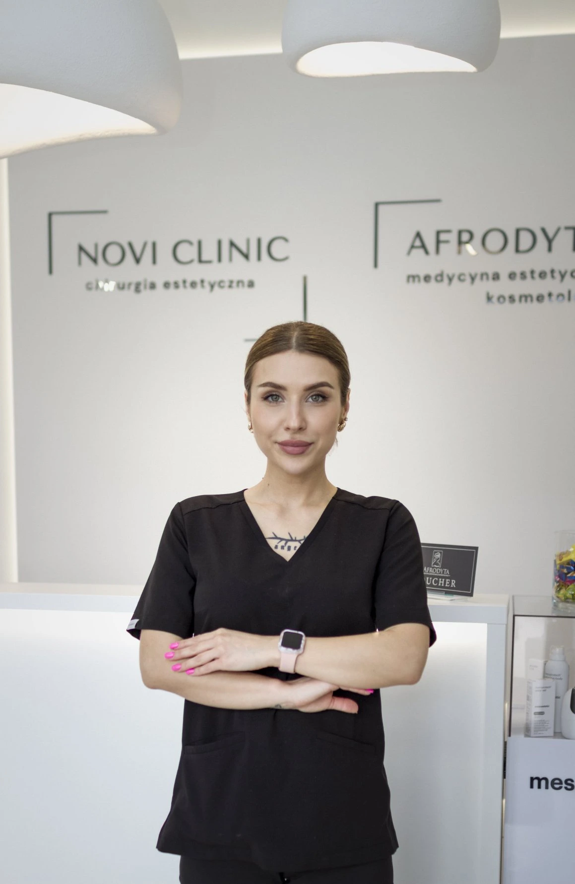 Kobieta w czarnym uniformie medycznym stoi z założonymi rękami w recepcji kliniki. W tle widoczne logotypy „Novi Clinic - chirurgia estetyczna” i „Afrodyta - medycyna estetyczna kosmetologia”.