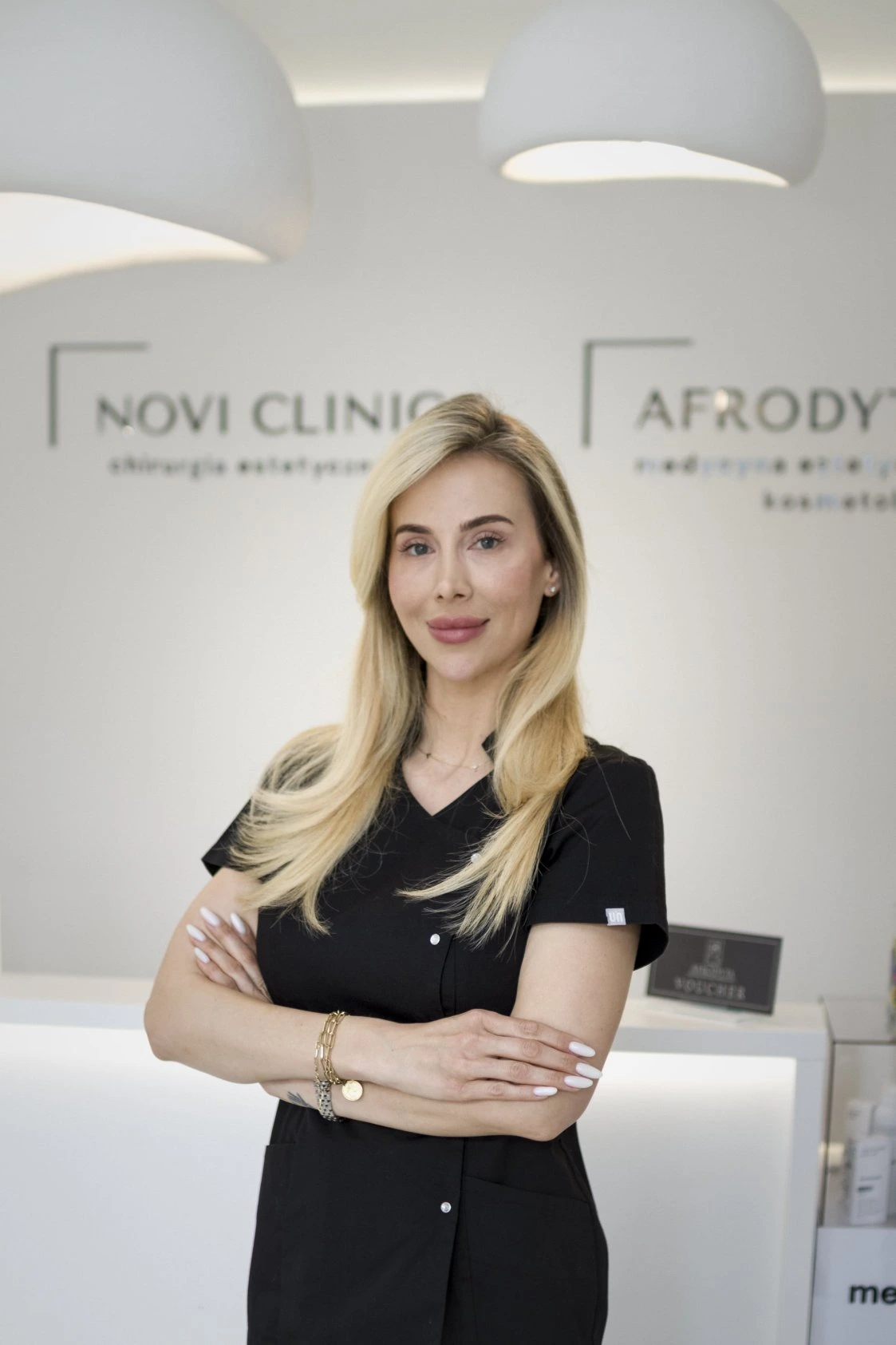 Portret trycholog Aleksandry Dworakowskiej w czarnym uniformie, stojącej z założonymi rękami na tle logotypów "Novi Clinic" i "Afrodyta".