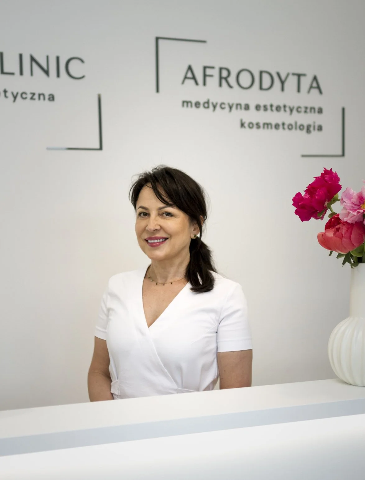 Portret Ewy Witek-Nowickiej w białym uniformie, stojącej za ladą recepcyjną kliniki. W tle widoczne logo "Afrodyta - Medycyna Estetyczna Kosmetologia" oraz wazon z kwiatami.