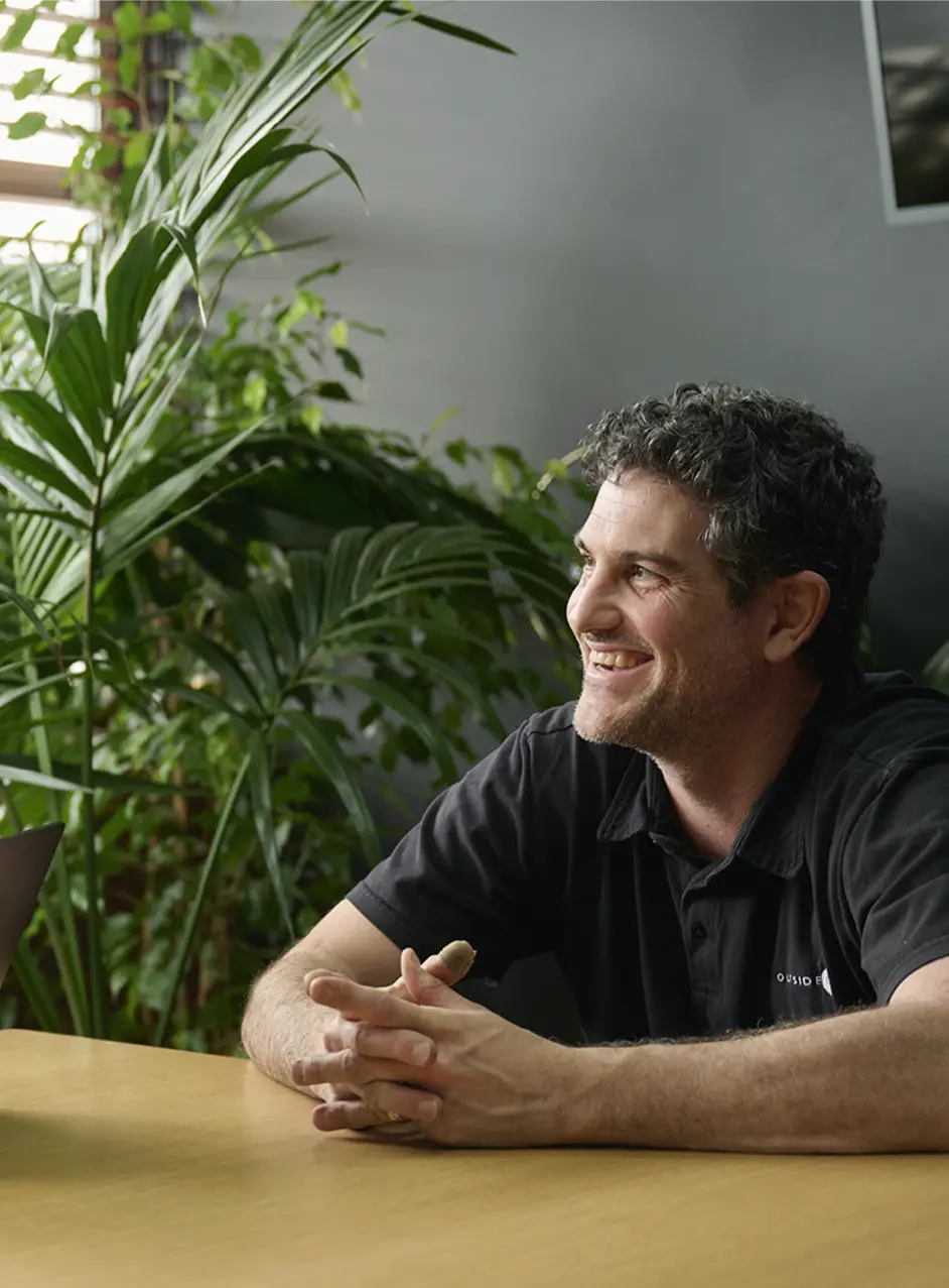 Smiling man with curly hair wearing a black polo shirt sitting at a wooden table with folded hands in a room with large green plants.