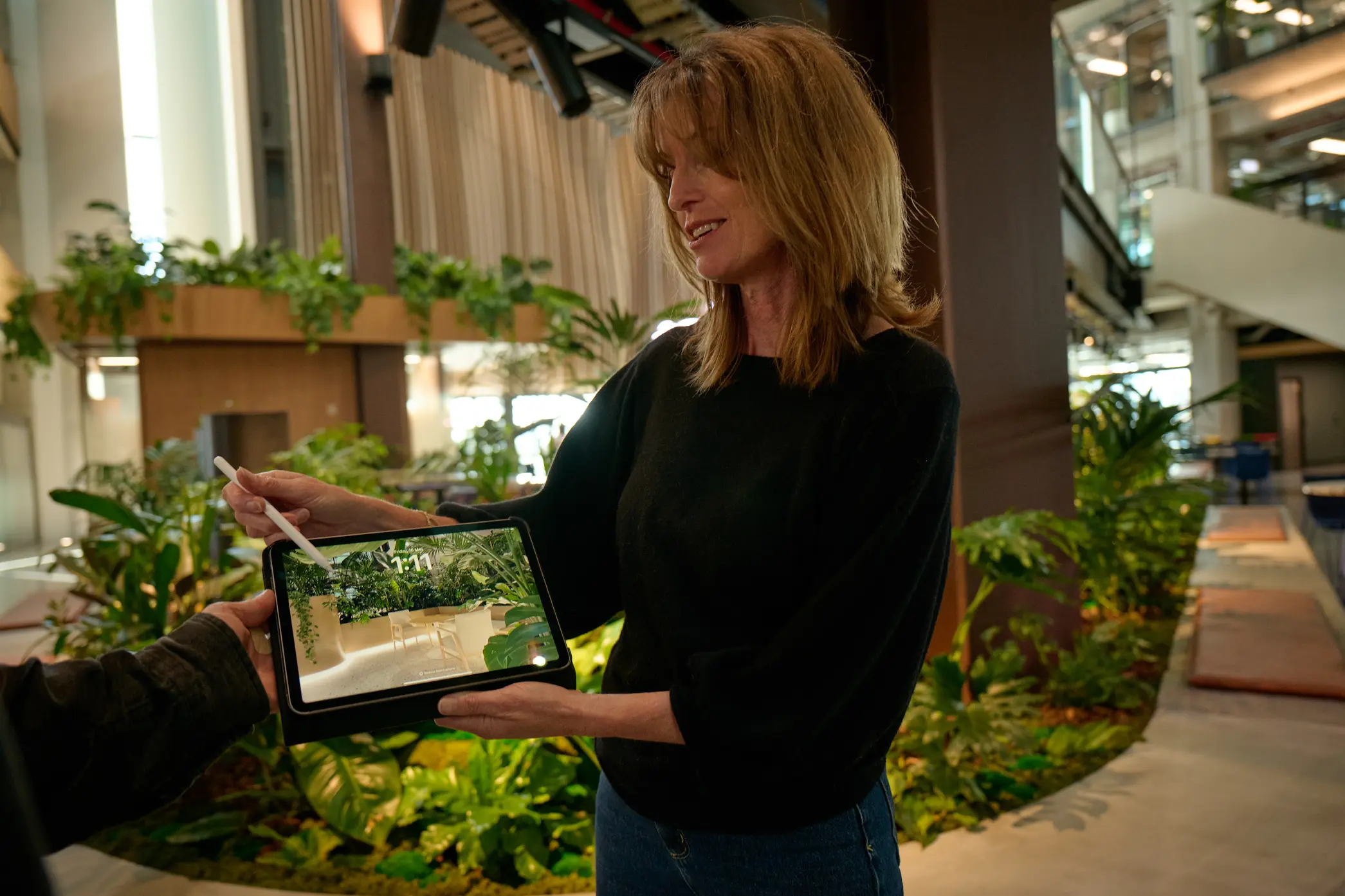 Woman holding a tablet displaying an interior design image of a room with plants and a chair.