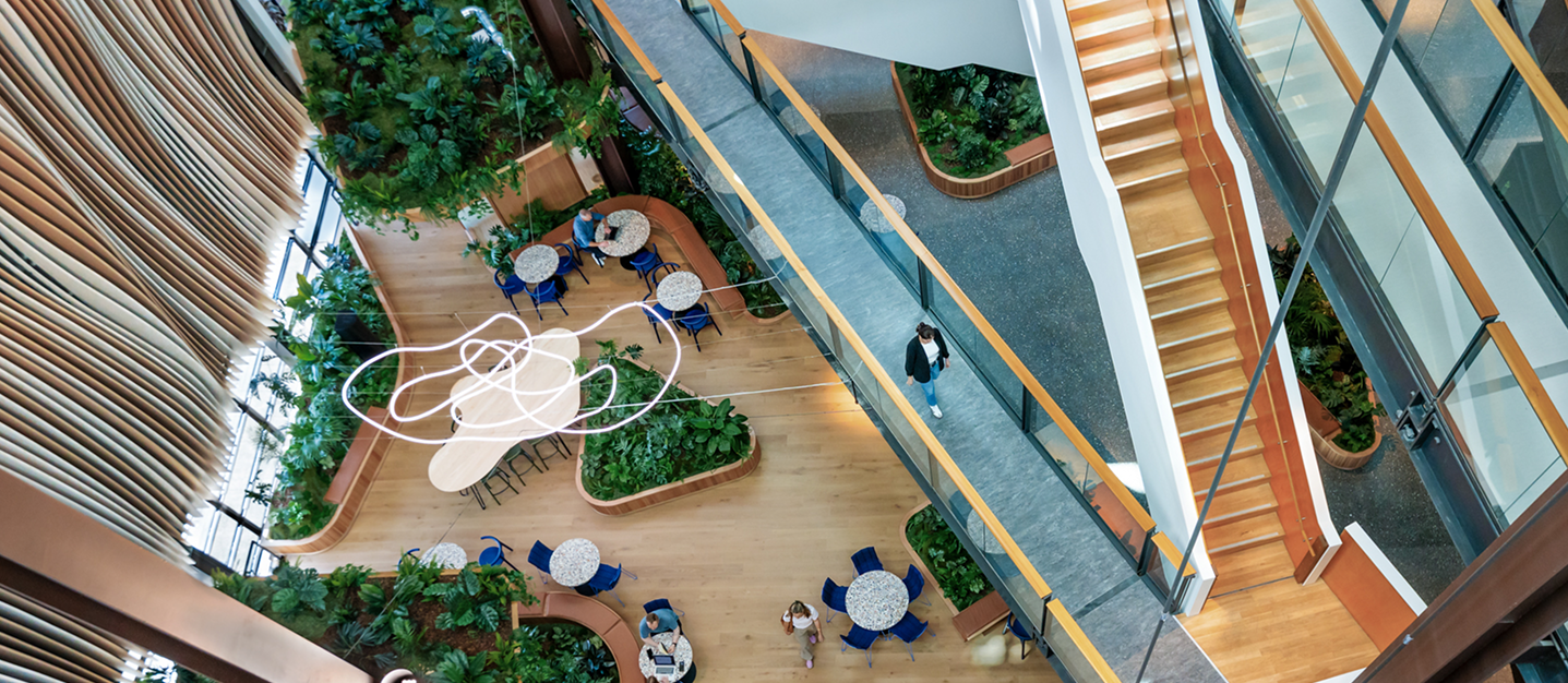Aerial view of a modern indoor space with wooden flooring, multiple round tables with blue chairs, green plants in planters, large windows with wooden blinds, and a person walking on a corridor beside a staircase.