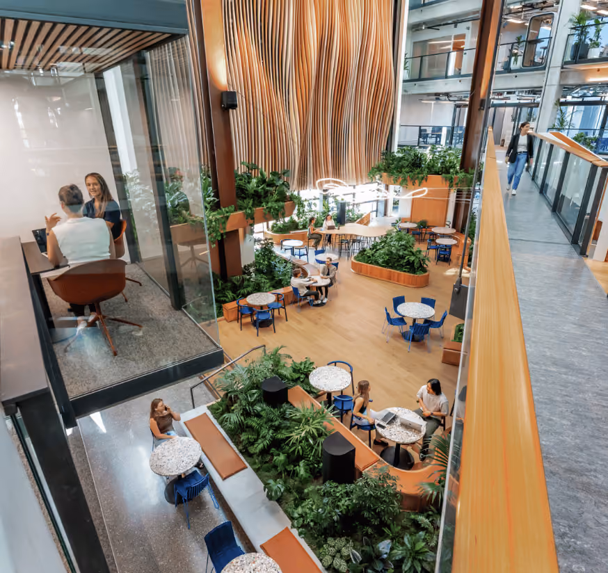Modern indoor office atrium with wooden floors, green plants, round tables, blue chairs, and people working and talking in a bright, spacious environment.