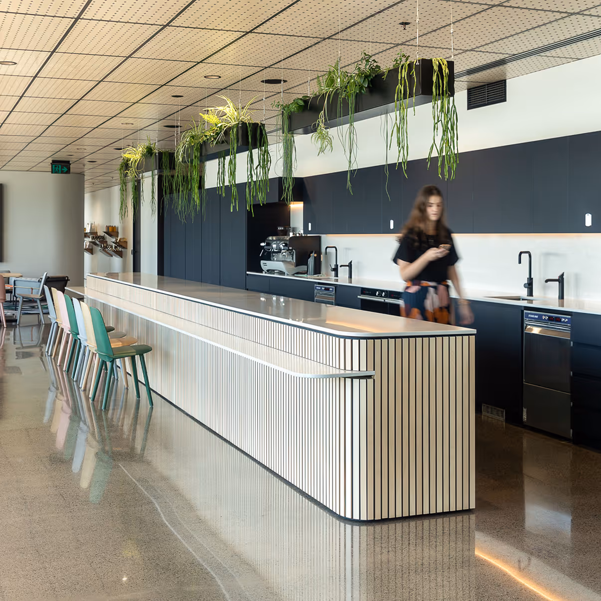 Modern office kitchen with a long striped island, colorful chairs, hanging plants, and a woman using a phone.