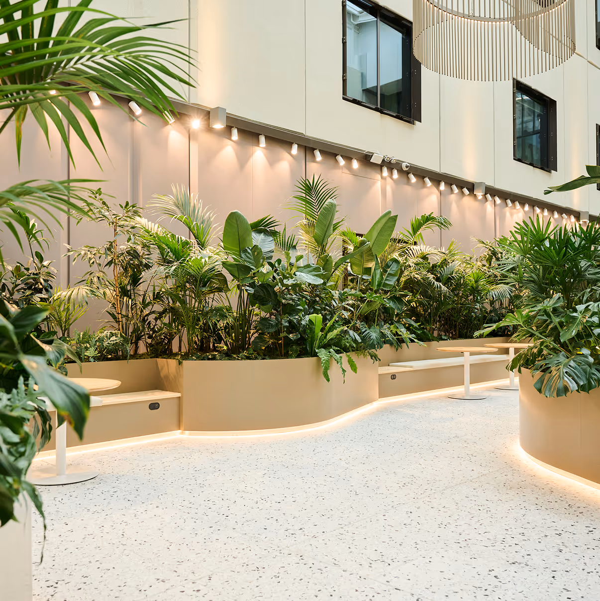 Indoor seating area with built-in benches and round tables surrounded by large green tropical plants under warm wall spotlights.
