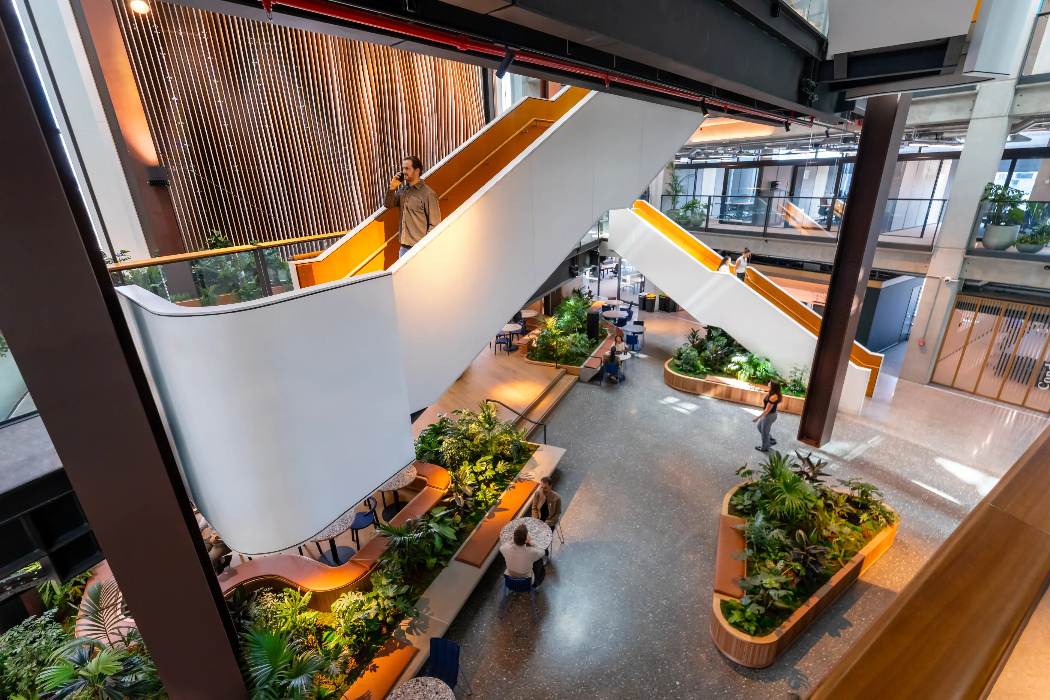 Side-by-side image showing a modern open-plan cafeteria before and after adding large white planters filled with lush green plants, brightening the space and adding natural décor near tables and seating areas.
