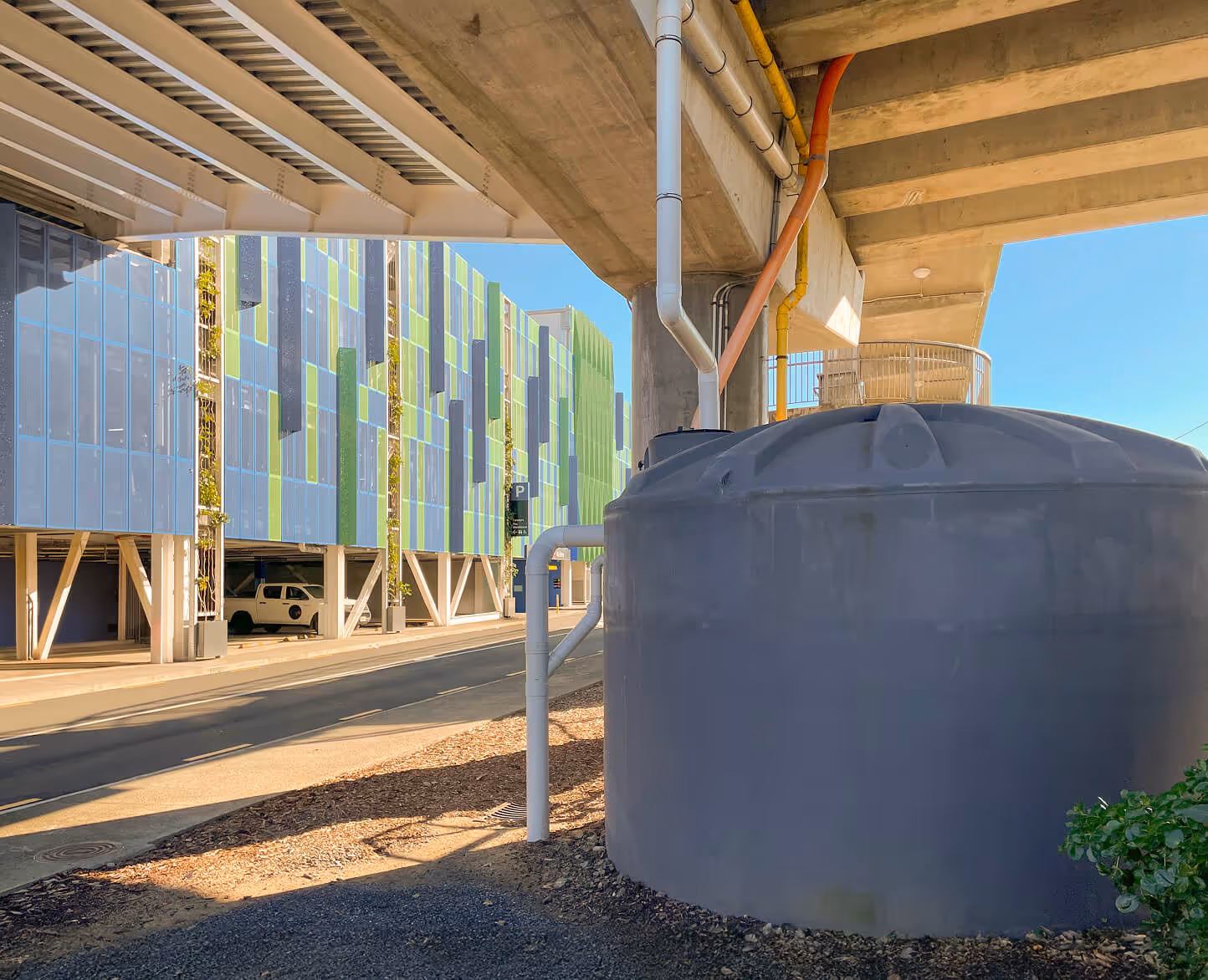 Large rainwater harvesting tank installed beneath an elevated transit structure next to a green-facade commercial building, showcasing sustainable water management design