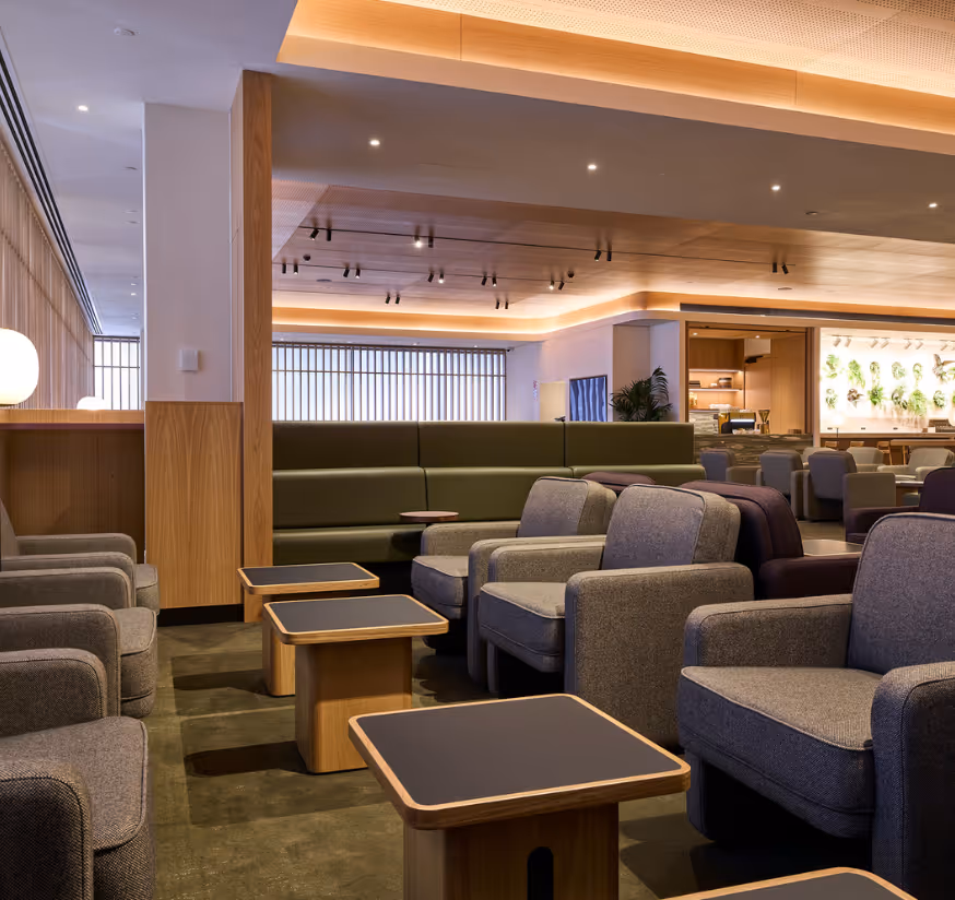 Open-plan seating area in the refurbished Qantas International Business Lounge, Auckland, with grey upholstered armchairs, timber coffee tables, and integrated planting creating a calm, biophilic retreat