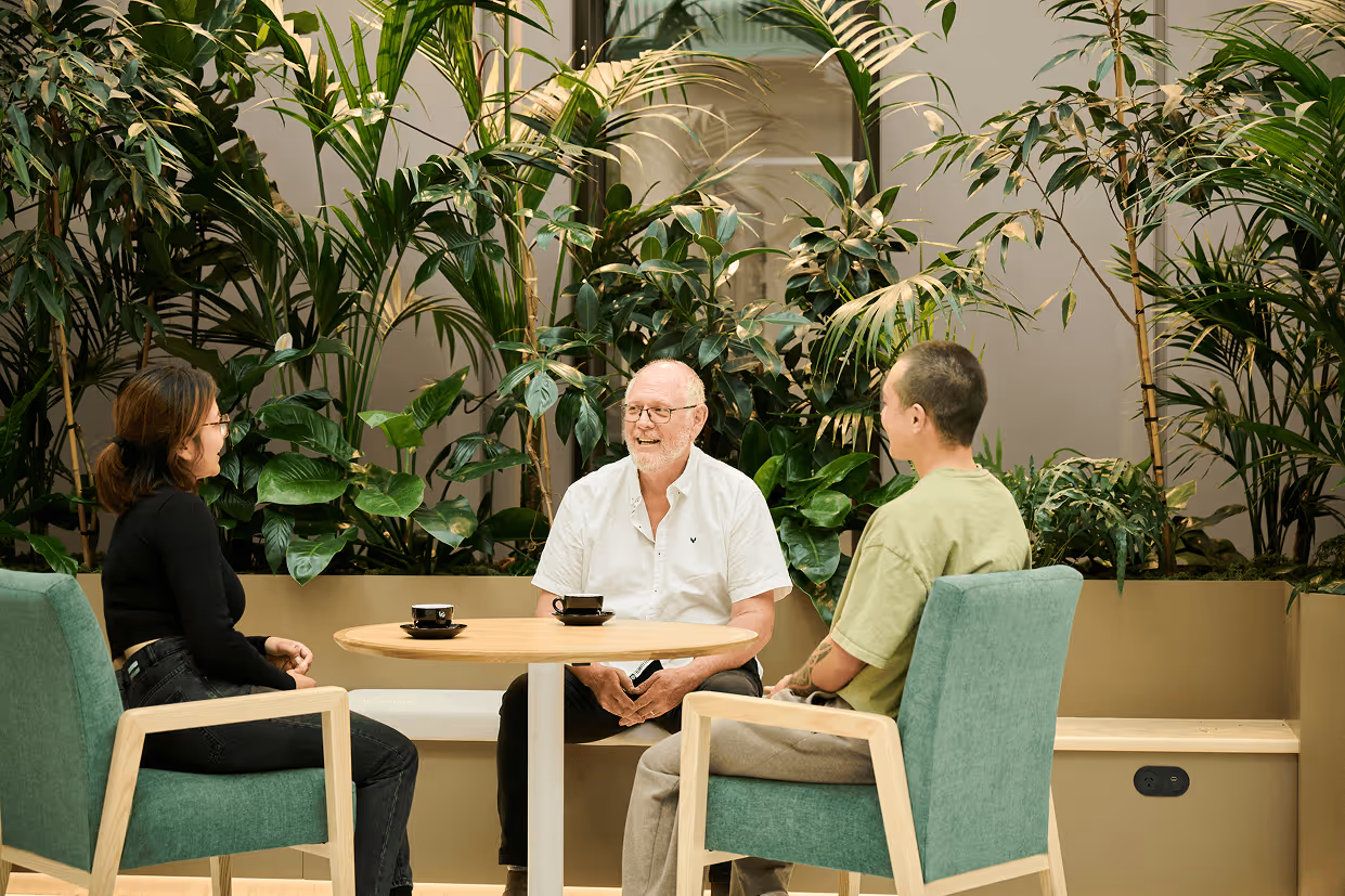 Two people having a relaxed conversation in a greenery-filled healthcare waiting area, showing how indoor plants create calmer, less clinical family spaces