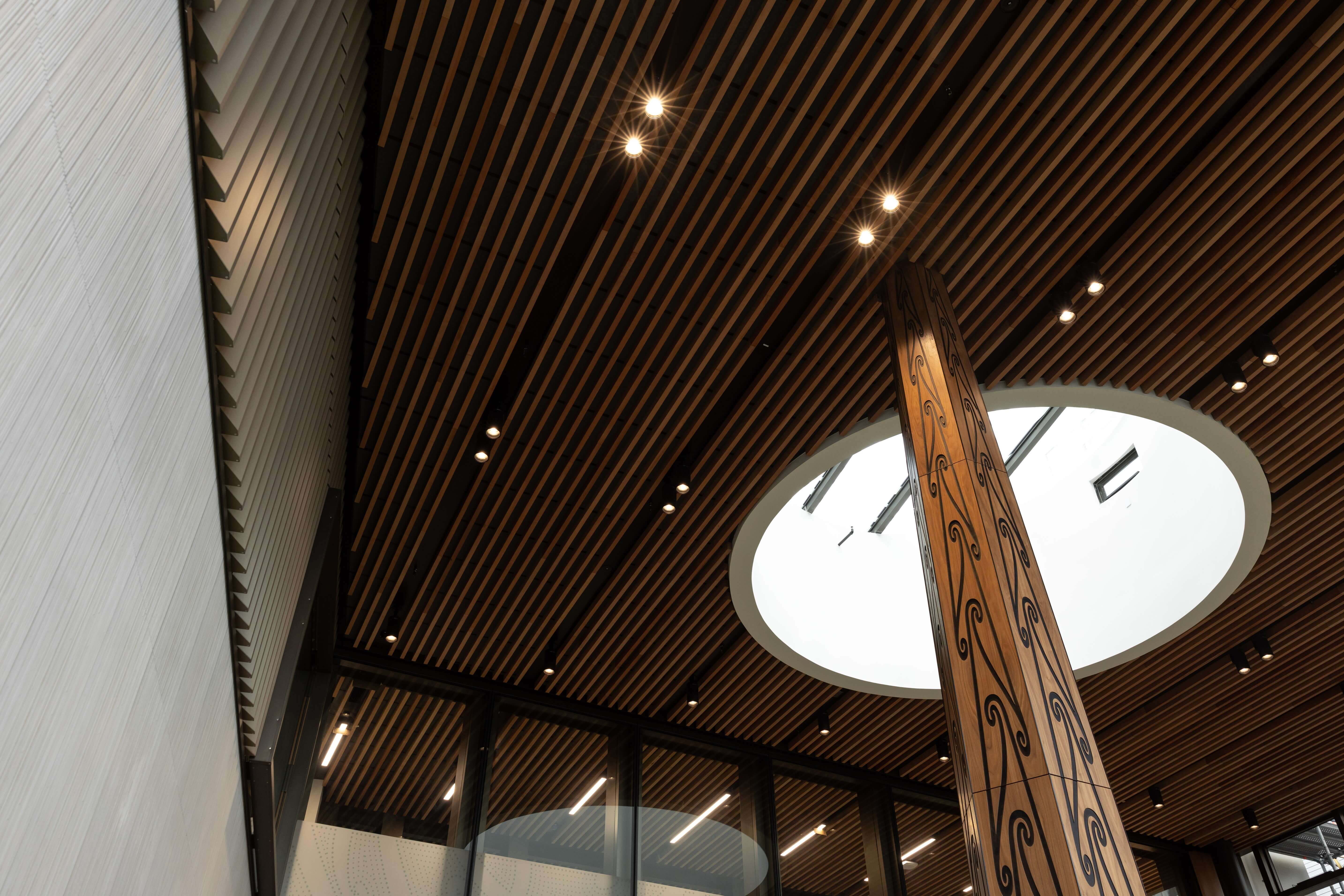 Carved timber pou rising through a circular skylight beneath a slatted timber ceiling.