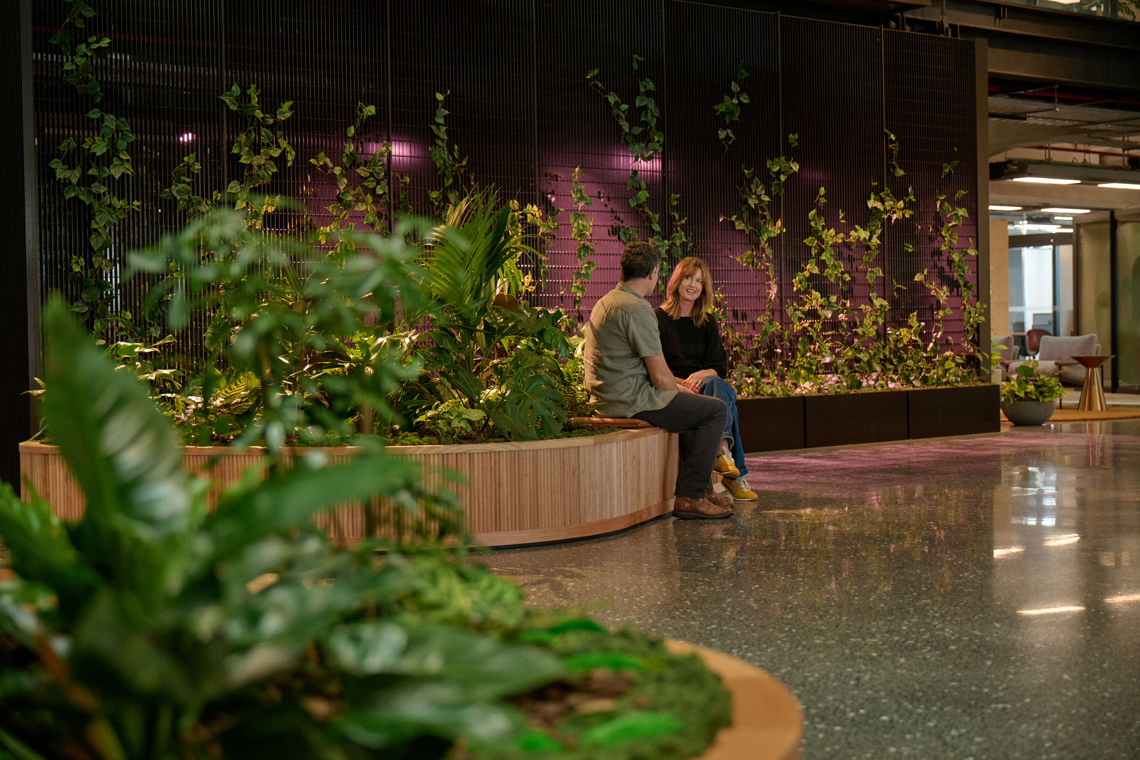 Colleagues sit beside a curved planter bench at B:HIVE — tropical greenery backed by a lit climbing plant wall.