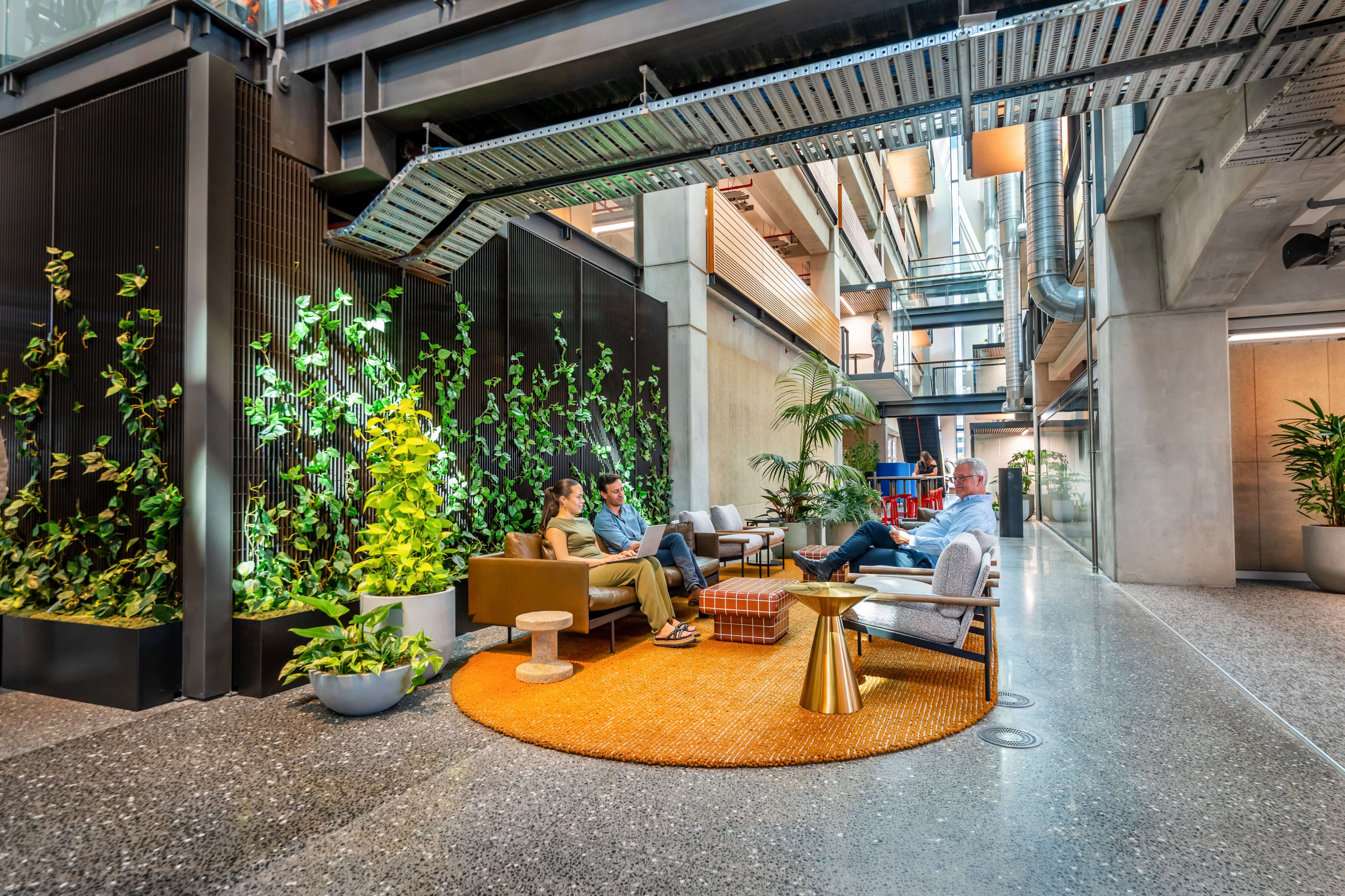 A pothos-covered vertical wall anchors a lounge area at B:HIVE — white bowl planters and an orange rug below the open atrium.