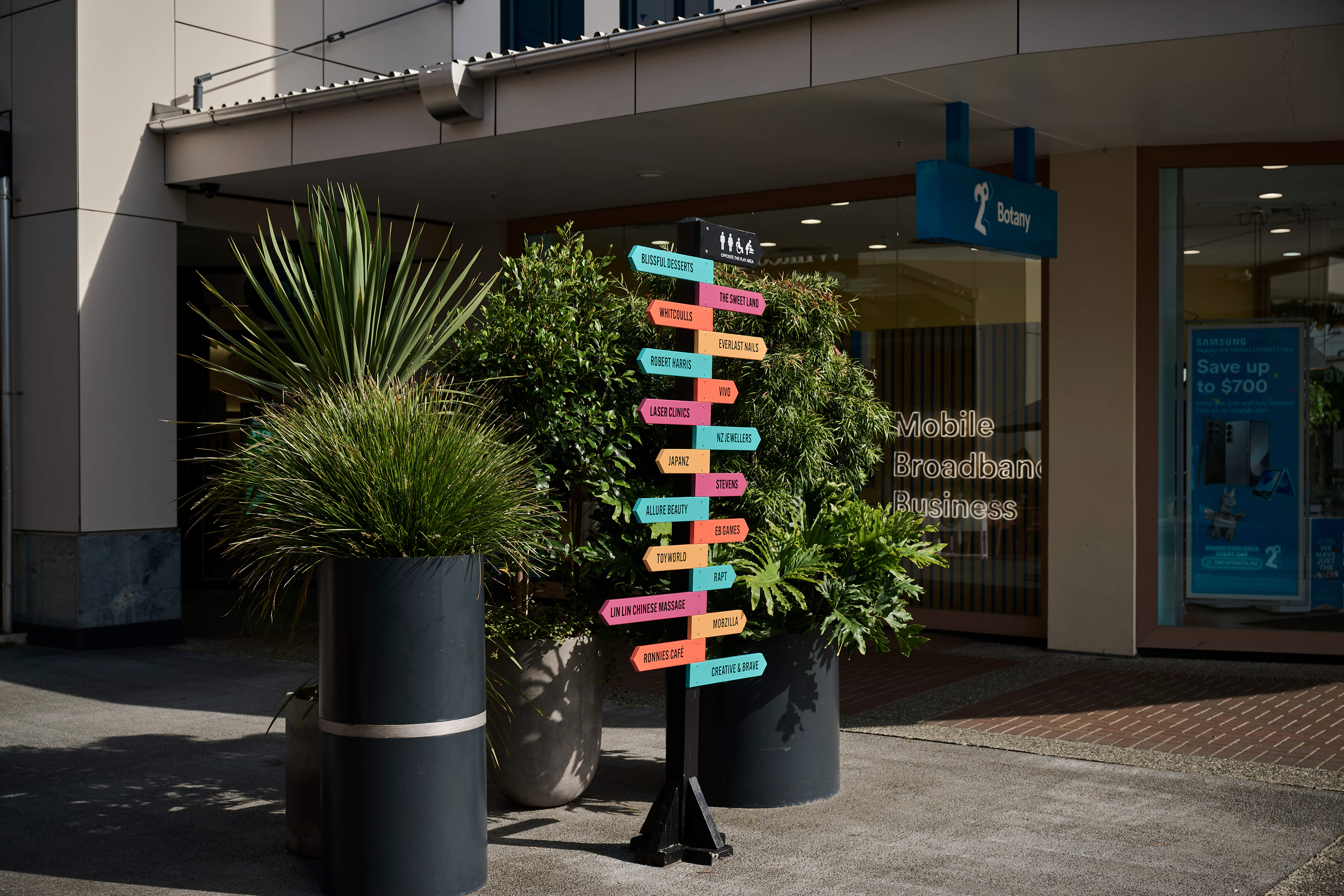 A colourful wayfinding sign flanked by cylindrical planters at Botany Town Centre's outdoor precinct.