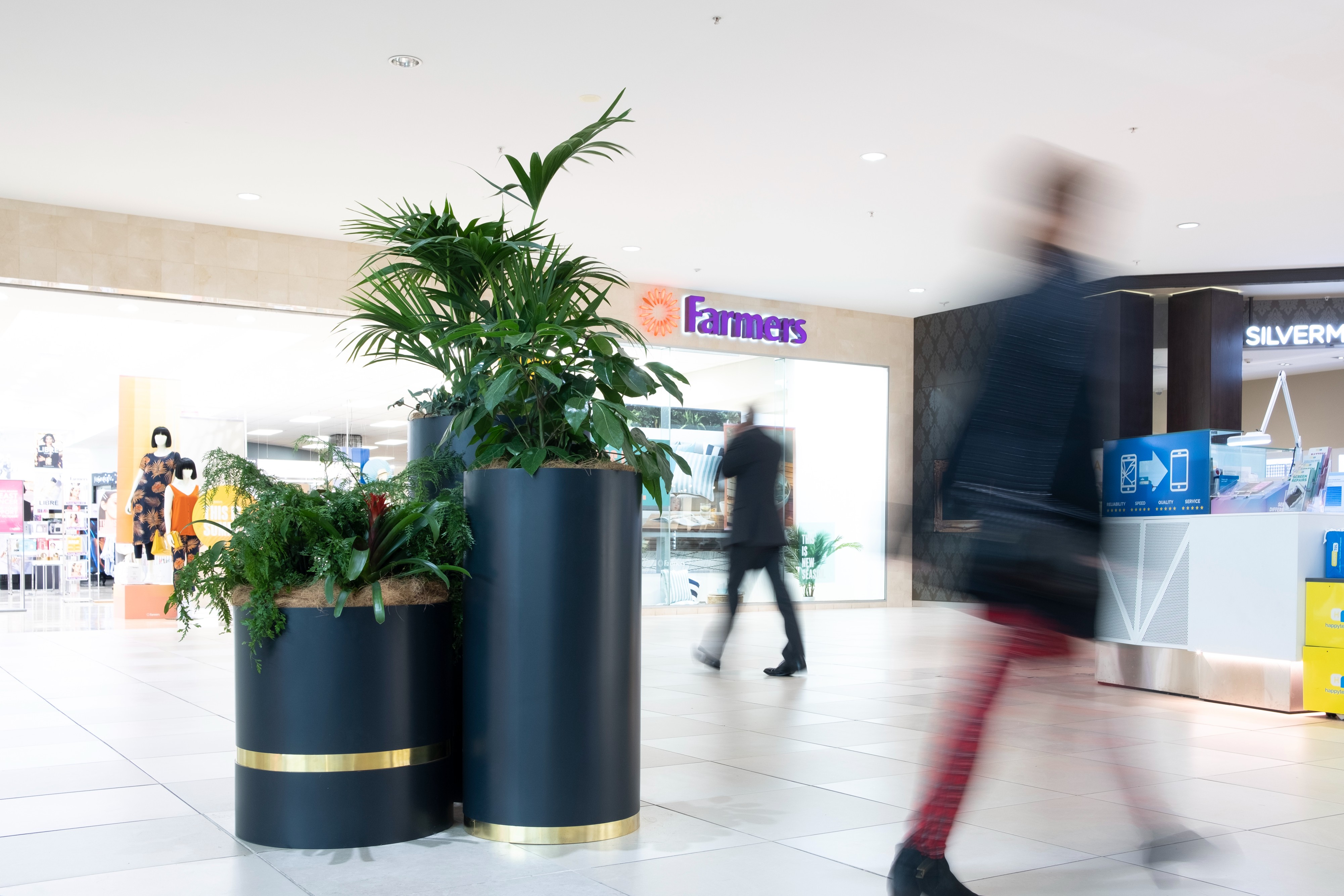 Shoppers move through Botany Town Centre's timber-arched arcade, with trailing ferns and tropical planters lining the.