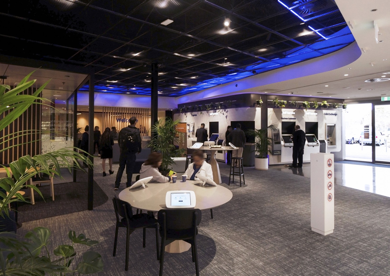 Bank branch floor with self-service kiosks, staff and customers, and potted palms beneath a blue-lit ceiling.