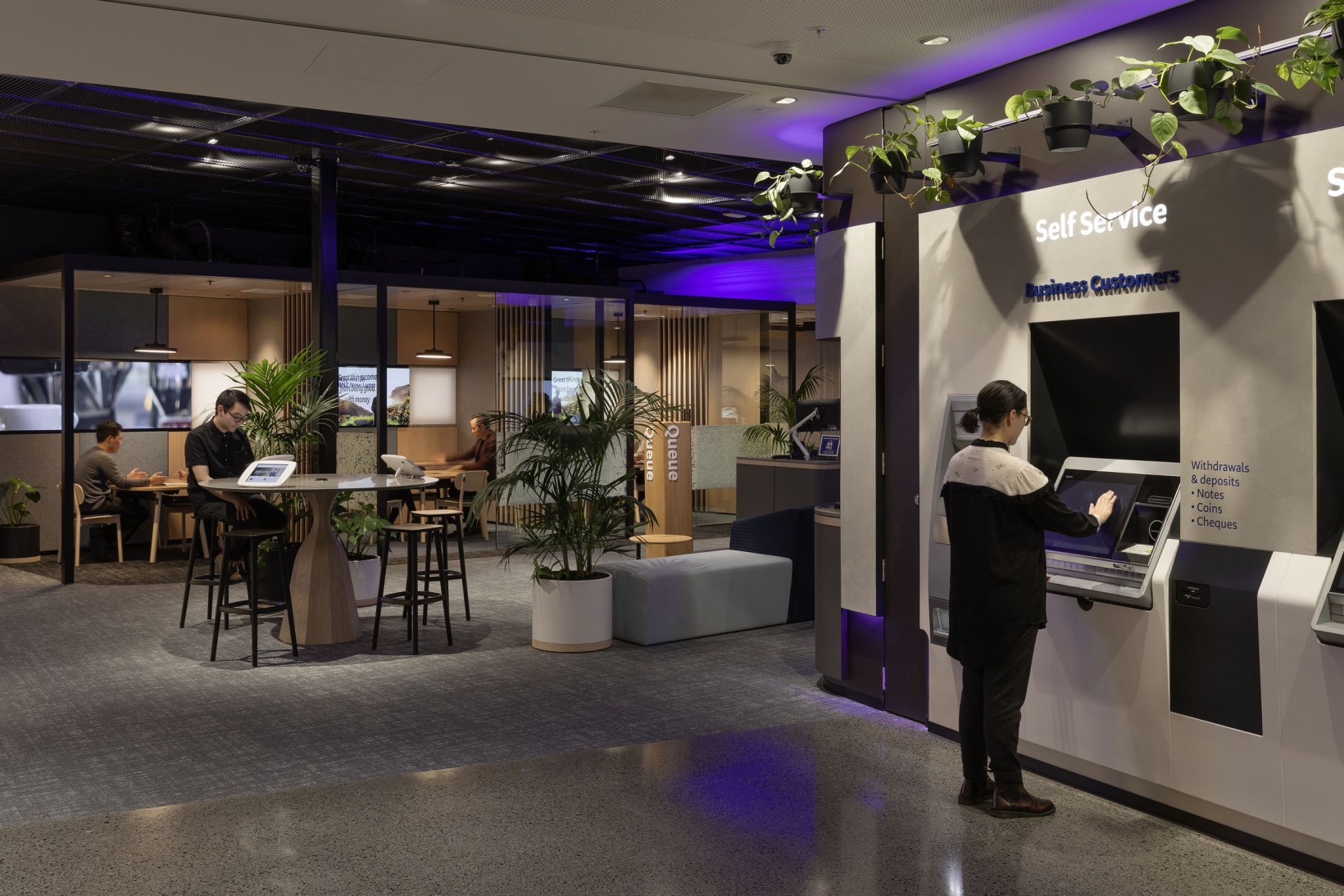 Bank self-service kiosk area with a customer at an ATM, palms and trailing pothos above the signage.