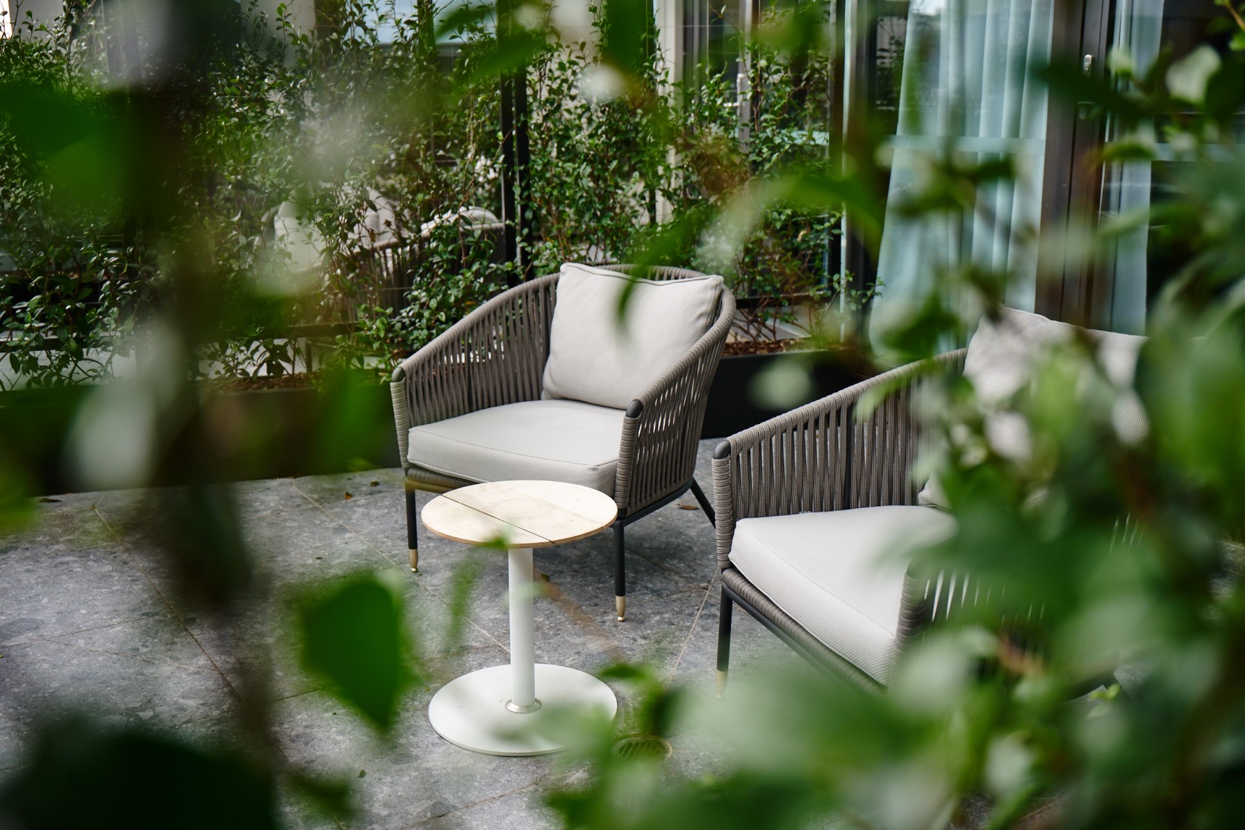 oven lounge chairs and a side table on an outdoor terrace, framed by dense foliage.