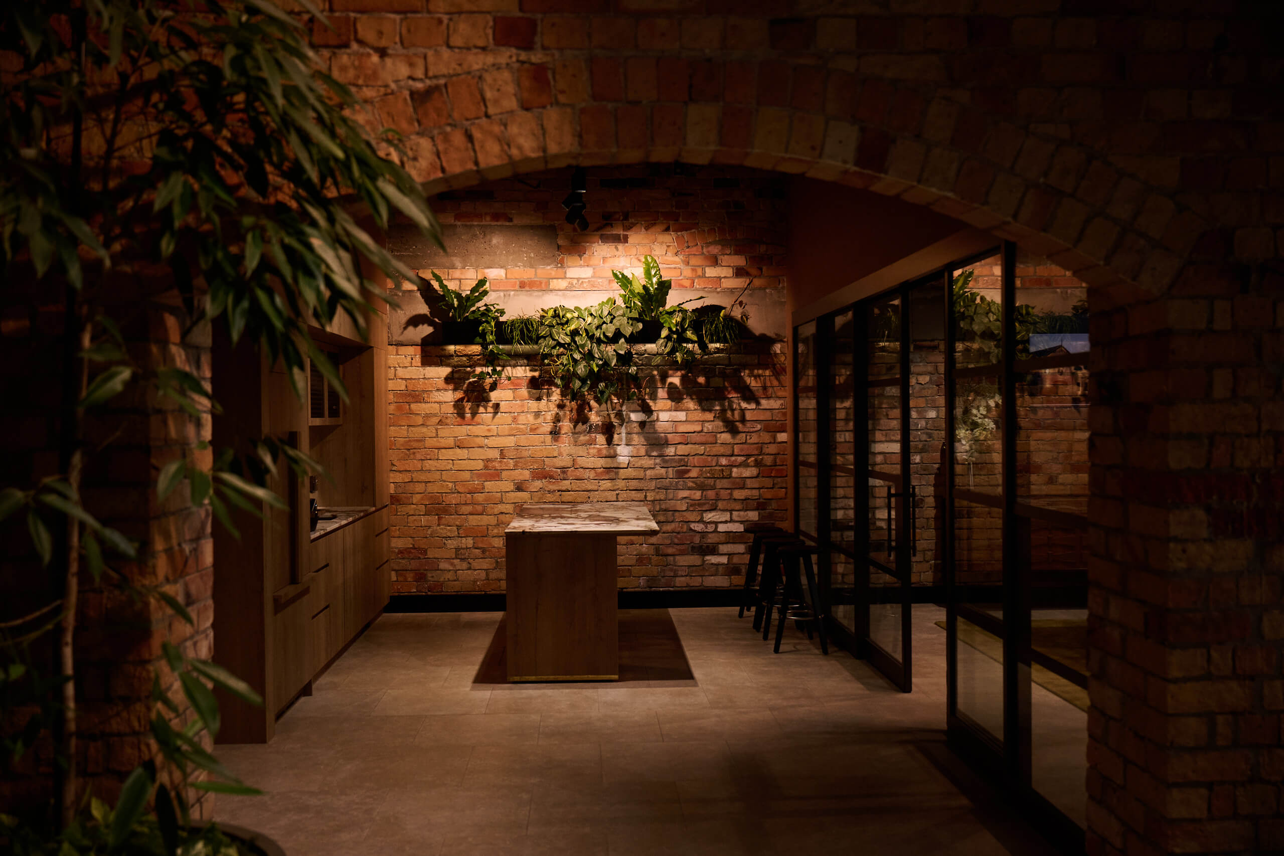 A bamboo planter frames the brick arch at Gold Merchants, with trailing pothos on a lit shelf above the marble kitchen.
