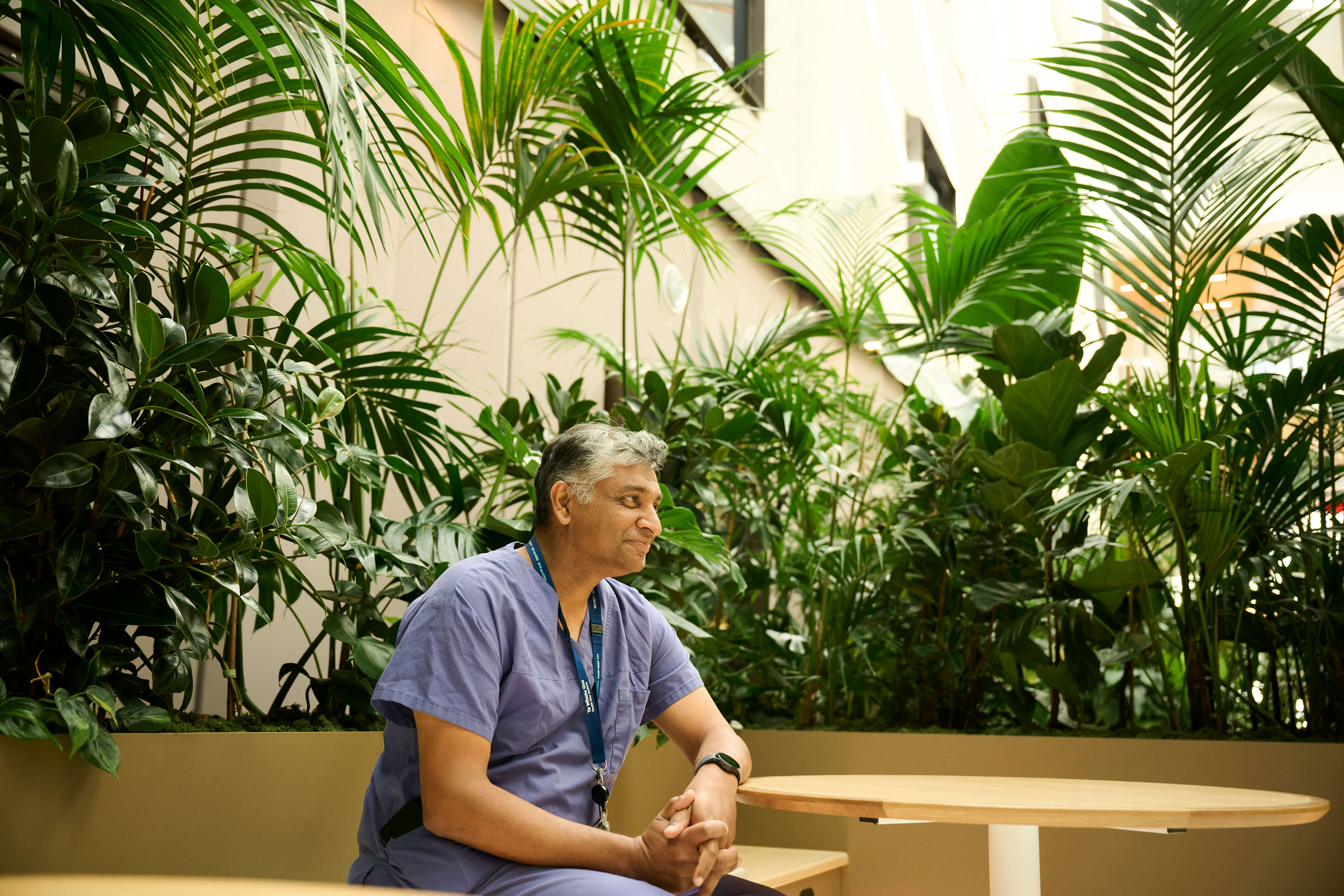 A clinician rests in the Totara Haumaru healing garden, surrounded by a canopy of palms, rubber plants, and tropical foliage.