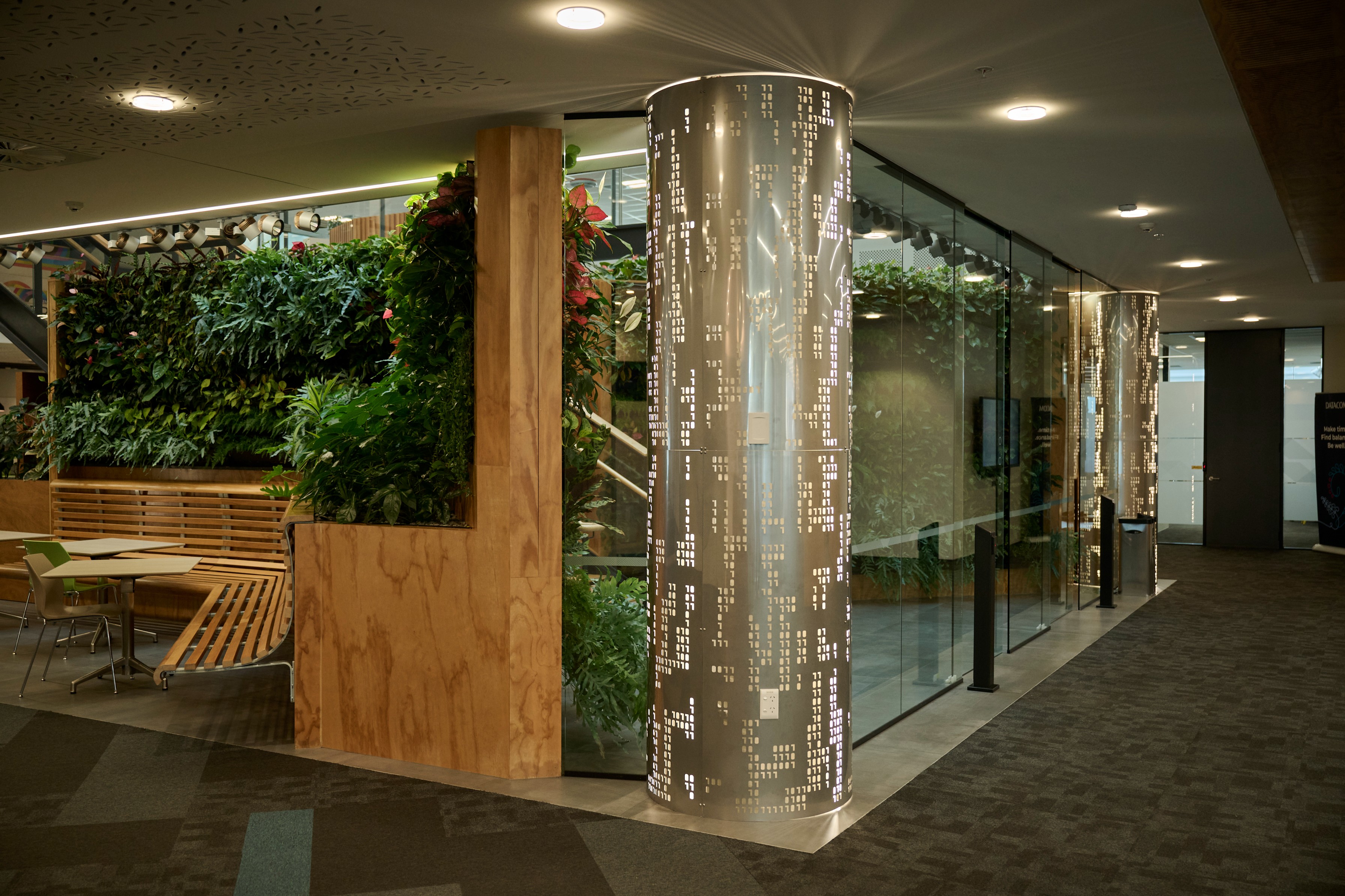 Green wall of ferns and anthurium framing timber booth seating beside a perforated metal column in an office corridor.