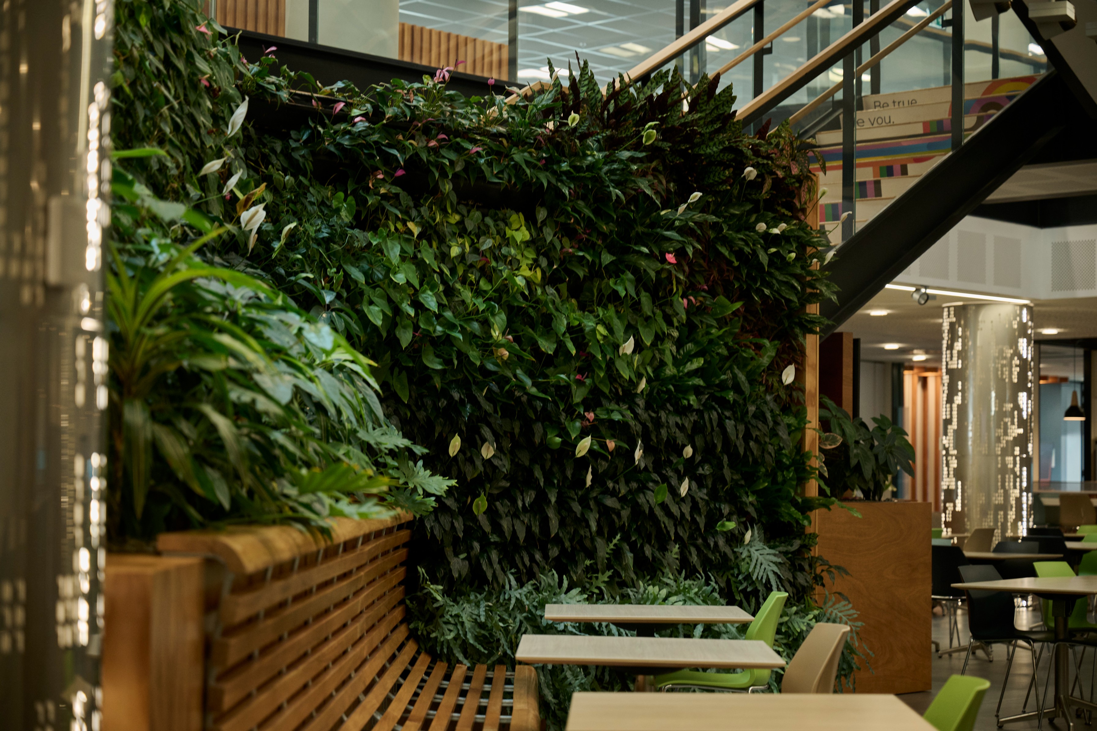 Flowering anthurium green wall curving beneath a staircase above timber-slat booth seating and cafe tables.