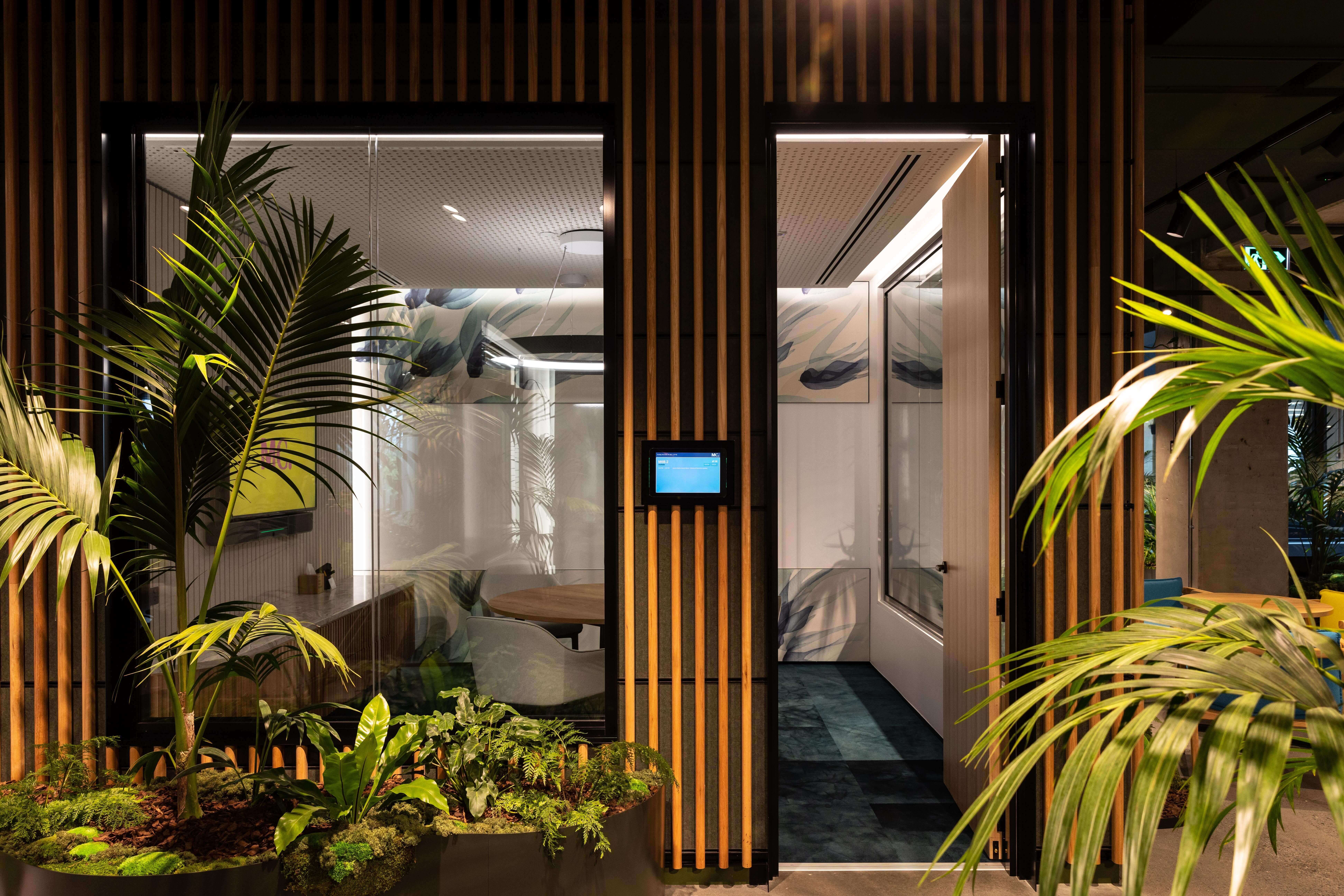 Glass-walled meeting pods at MC are surrounded by kentia palms and tropical planting — nature framing every workspace.