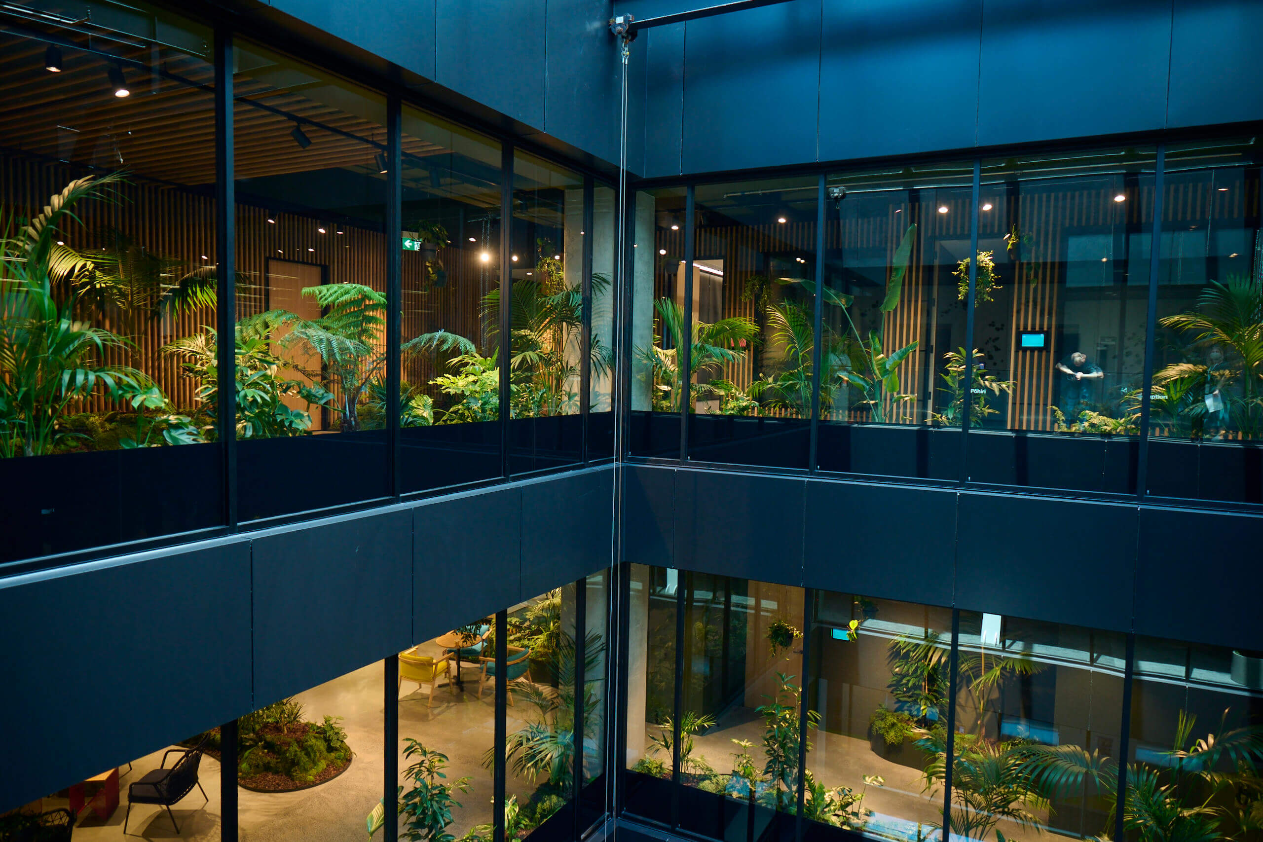 Tropical planting visible across two floors through dark-framed glass at MC — a fully immersive biophilic workplace.