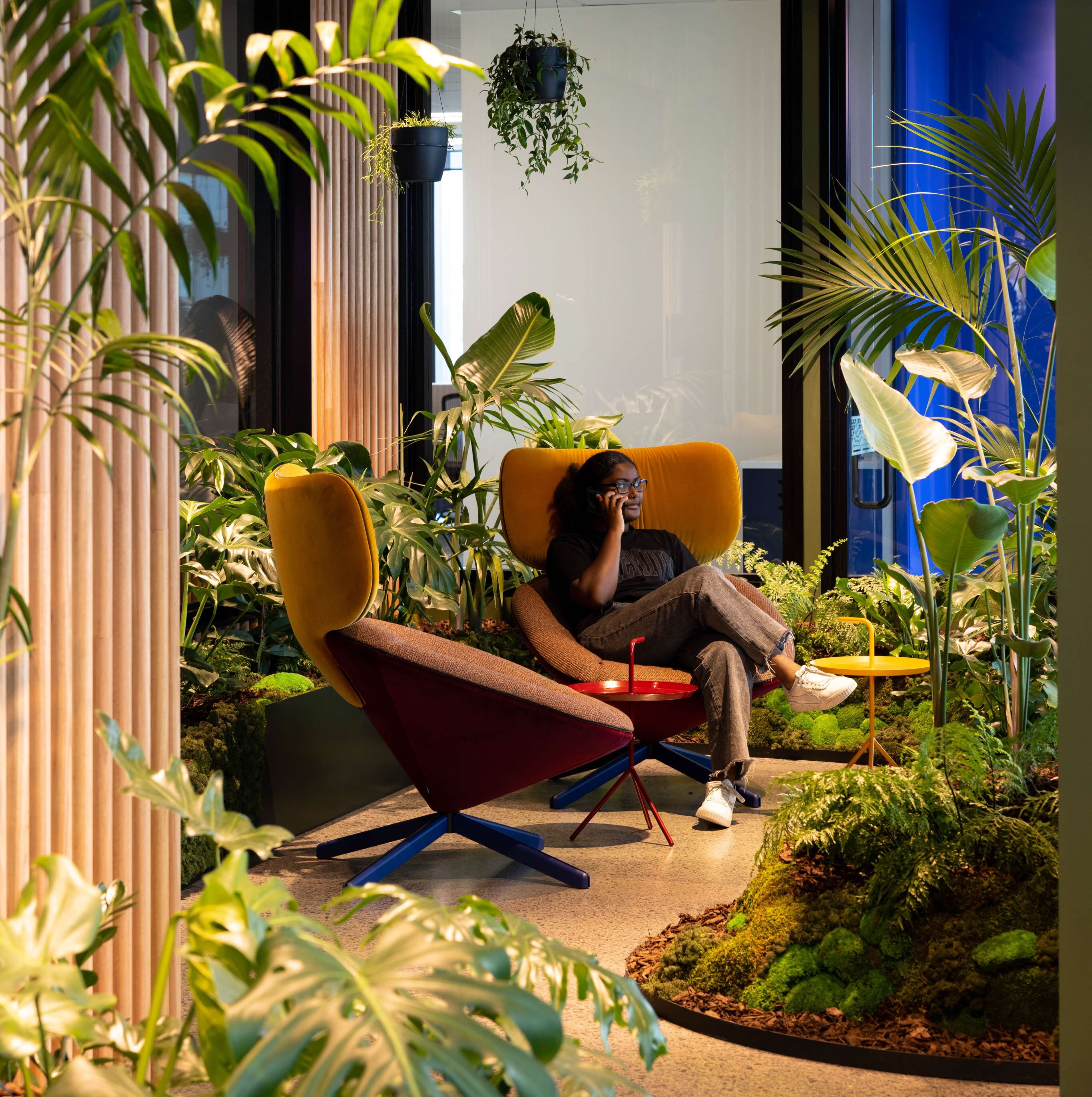 A person takes a call in a mustard lounge chair surrounded by moss, ferns, and tropical plants at MC.