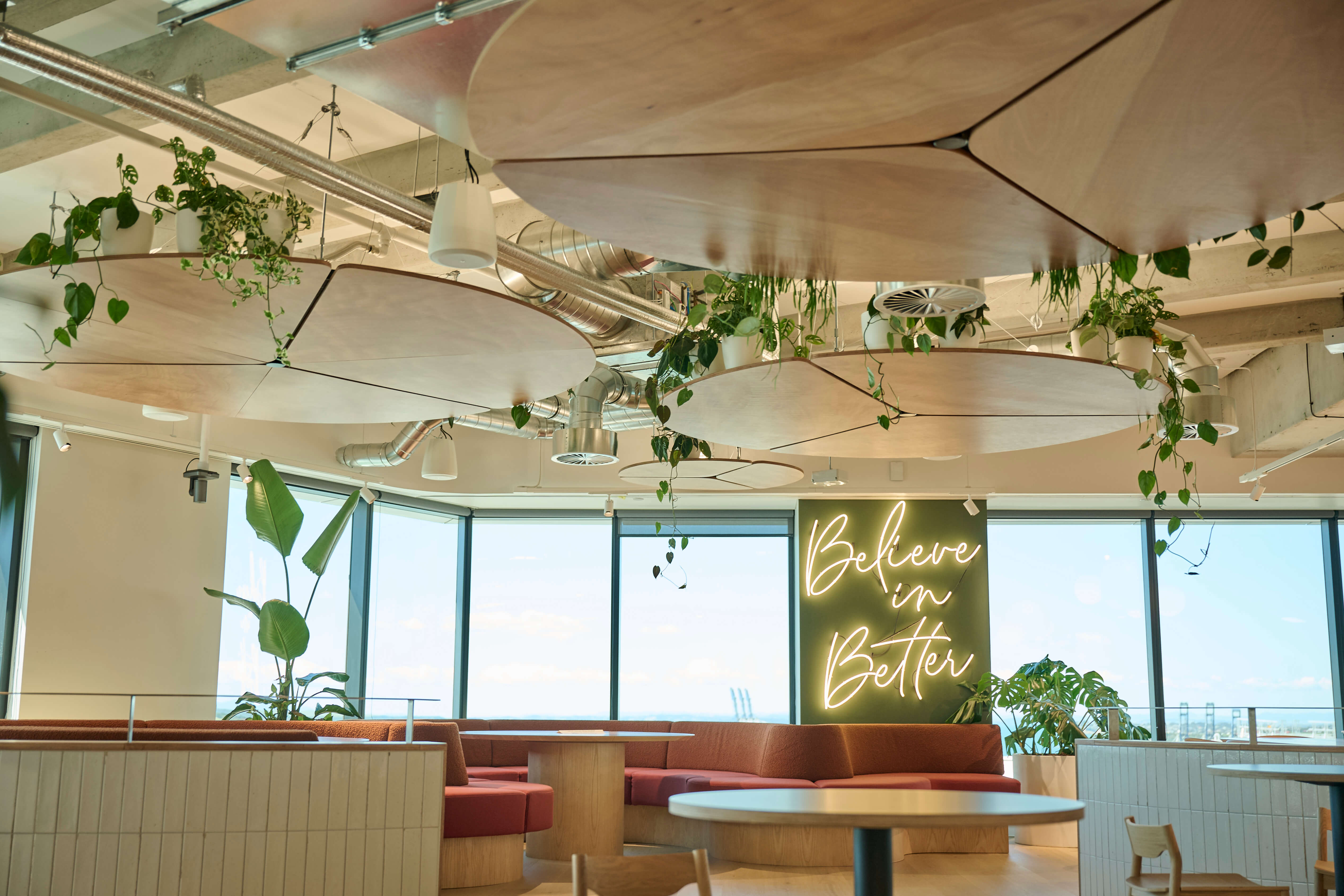 Plywood cloud baffles and hanging plants frame Oceania's lounge, with terracotta sofas and a neon 'Believe in Better' sign.