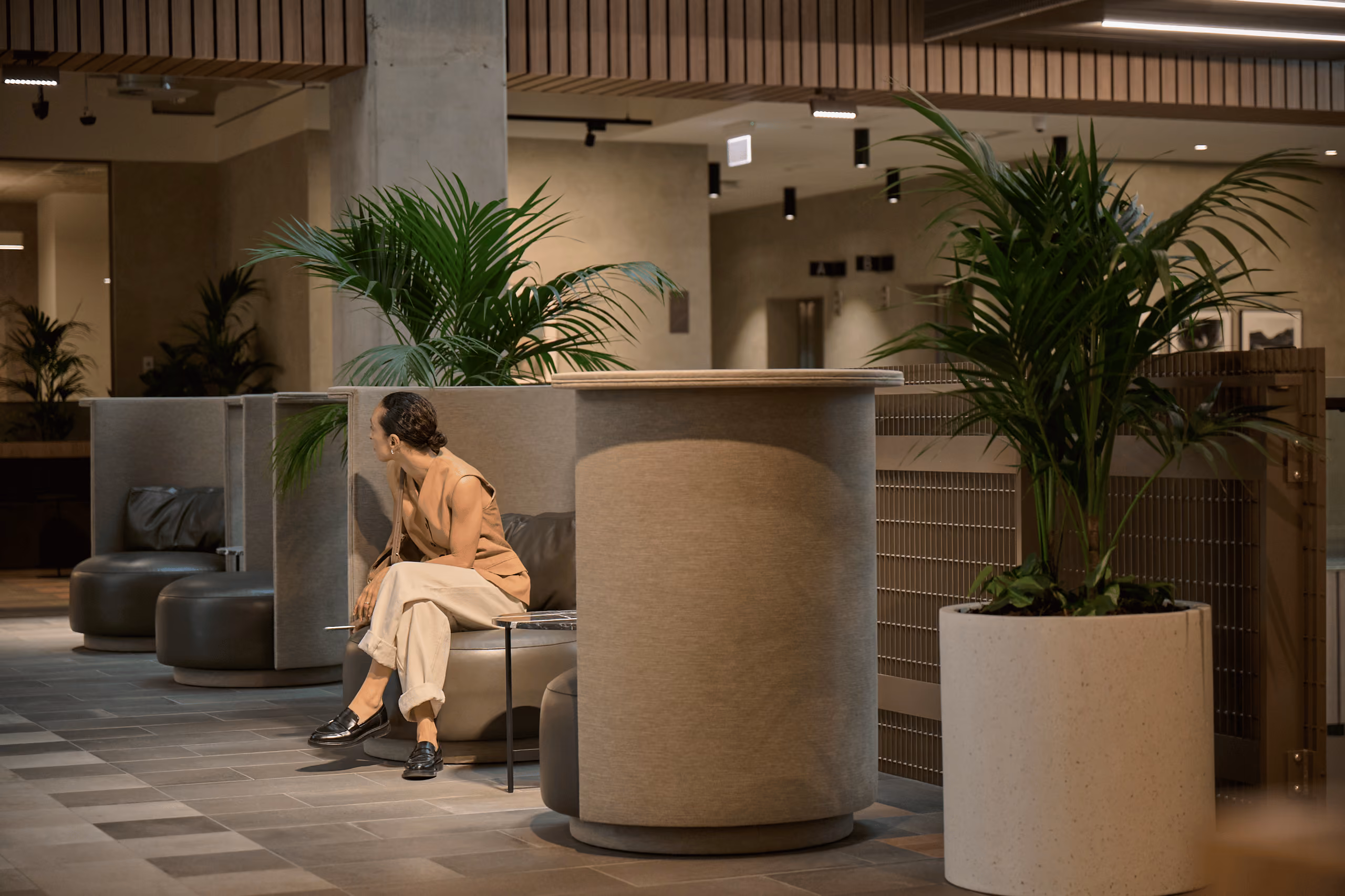 Kentia palms in tall planters beside pod seating and a woman seated in a lobby lounge area.