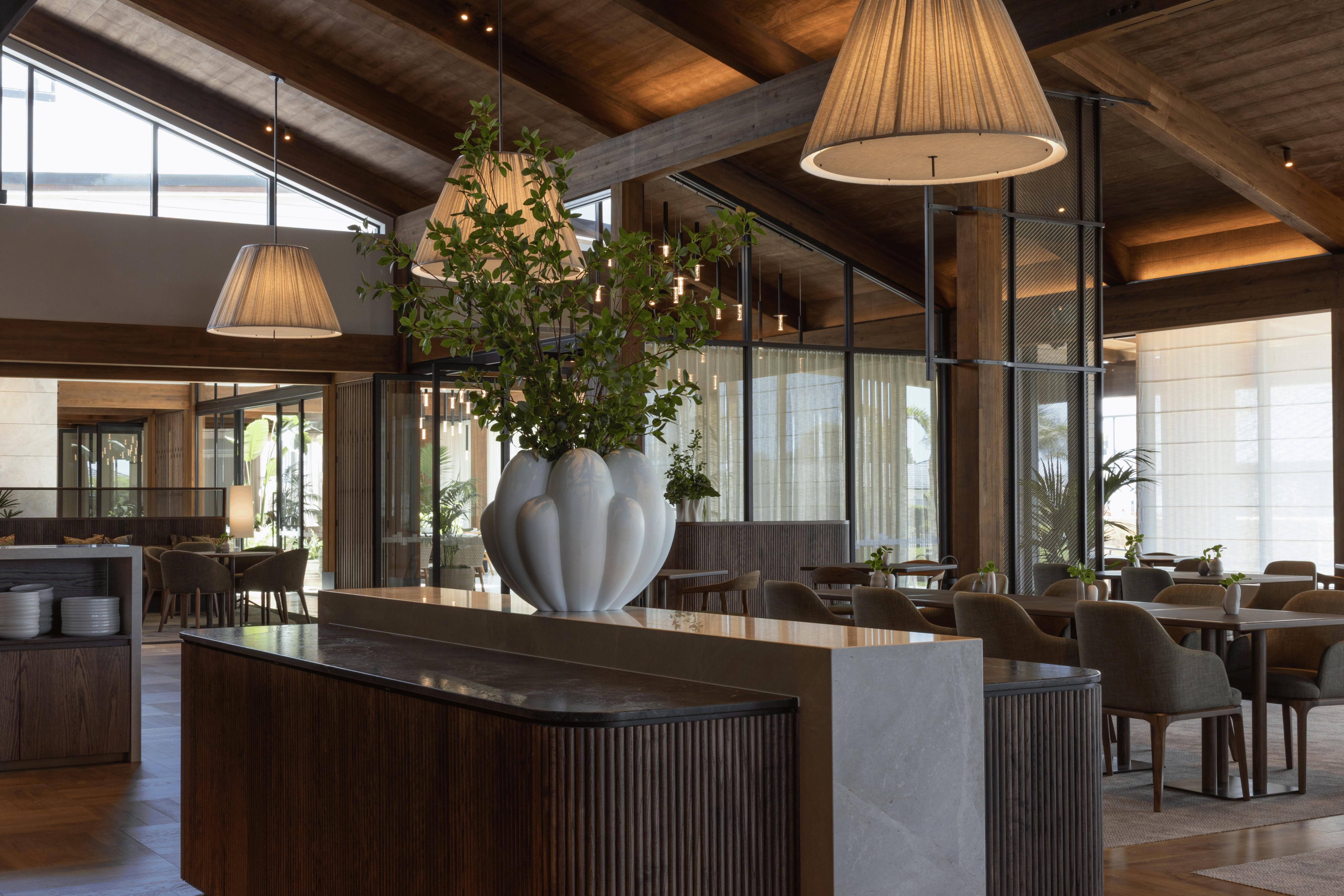 A large scalloped white vase of foliage on a stone-topped bar counter beneath pleated pendant lights in an open dining room.