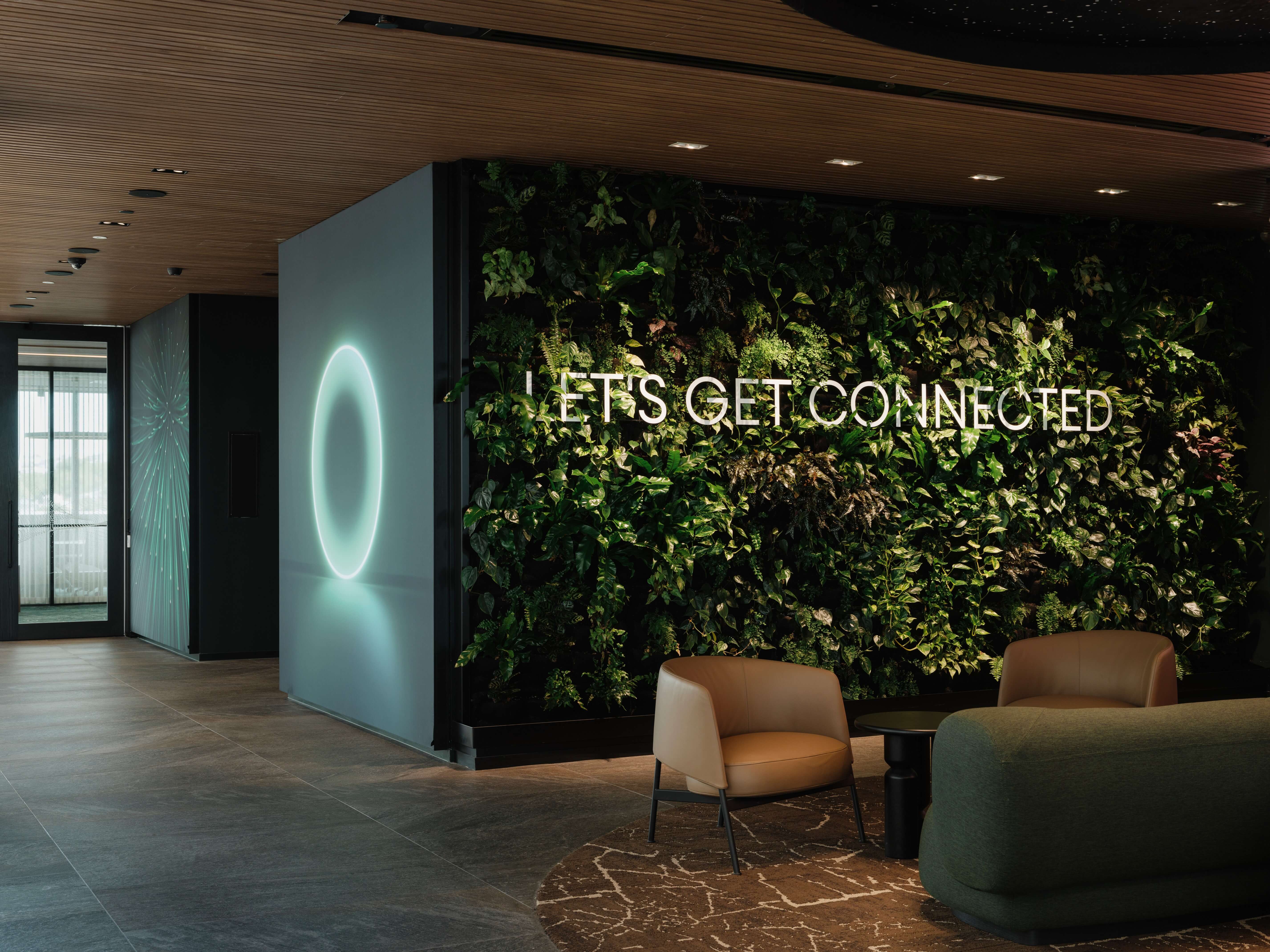 "Let's get connected" neon ring sign in front of a full-height green wall, with terracotta and olive lounge chairs on a patterned rug.