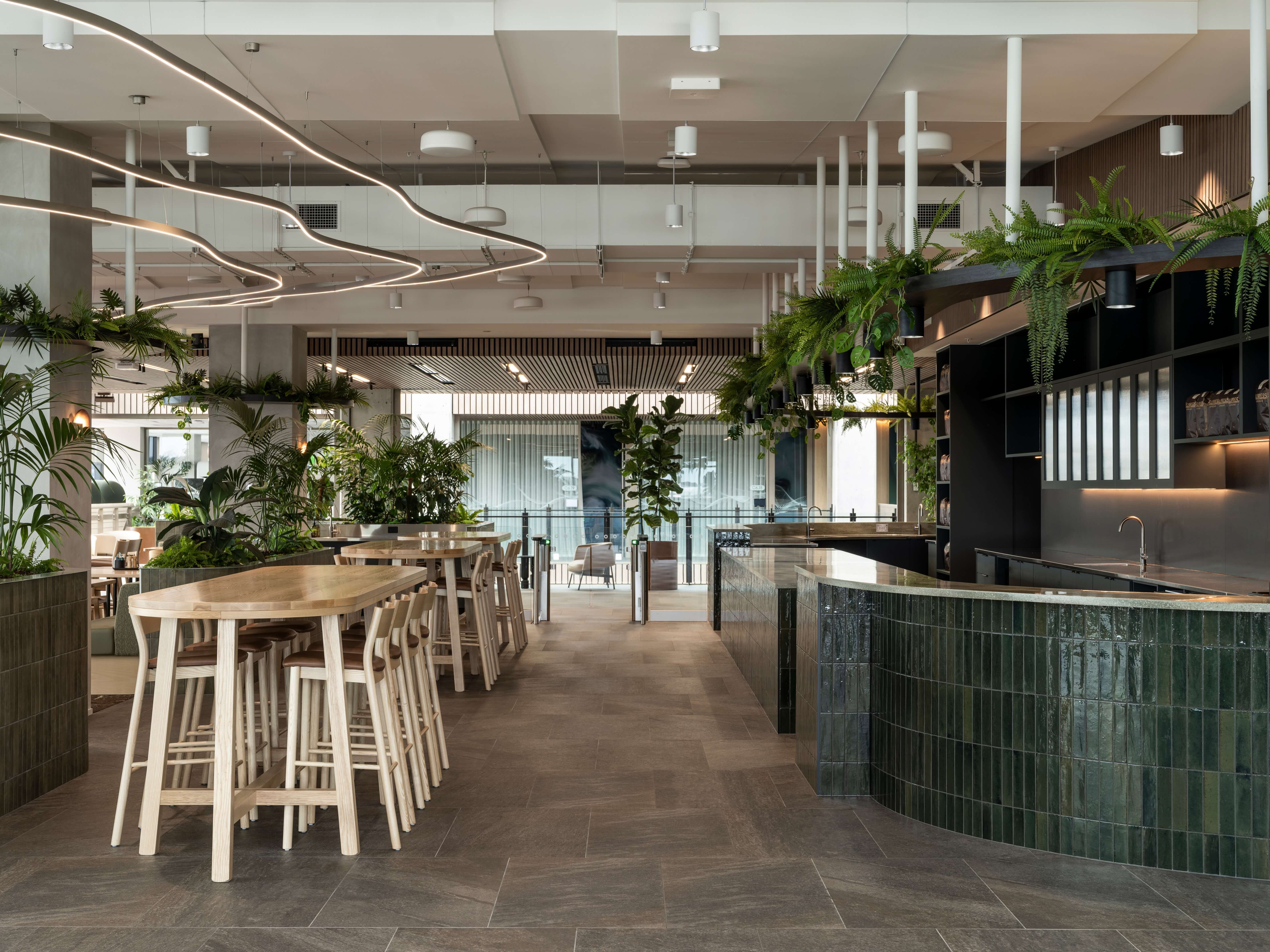 Oak café tables beneath organic curving LED lighting, surrounded by hanging ferns and tropical planting beside a dark-tiled kitchen counter.