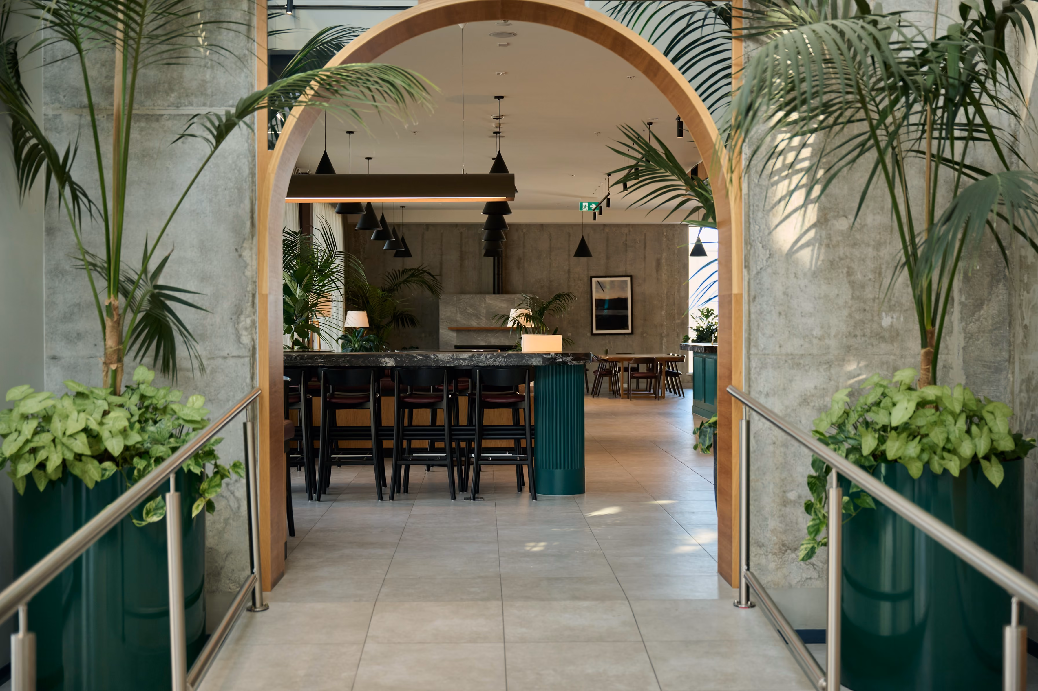 Green cylindrical planters with pothos and palms flanking the timber arch entrance to Sudima Auckland's bar and restaurant.