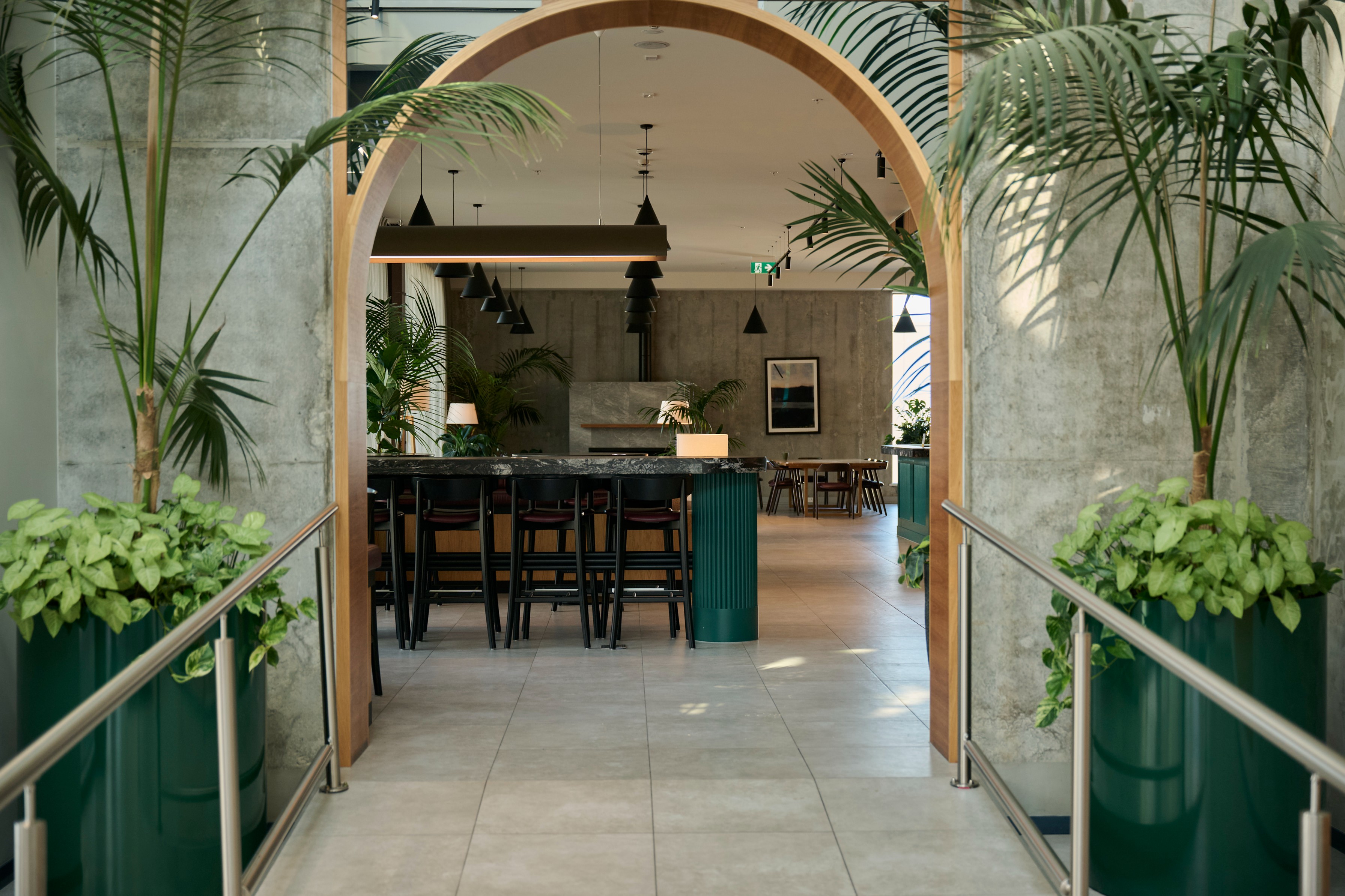 Green cylindrical planters with pothos and palms flanking the timber arch entrance to Sudima Auckland's bar and restaurant.