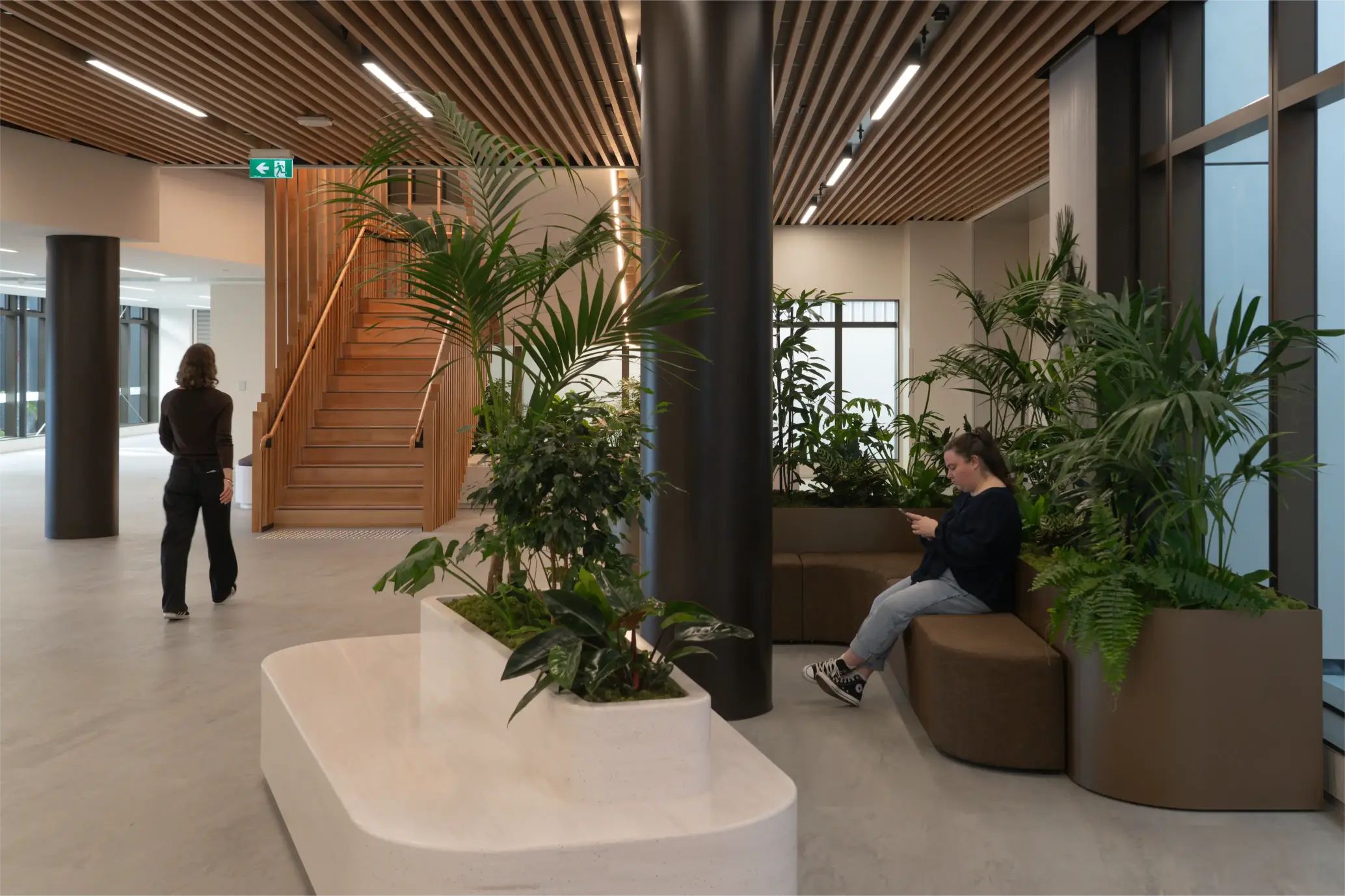 Hospital foyer with lush planter beds, curved seating, and a timber staircase, one person resting among the greenery