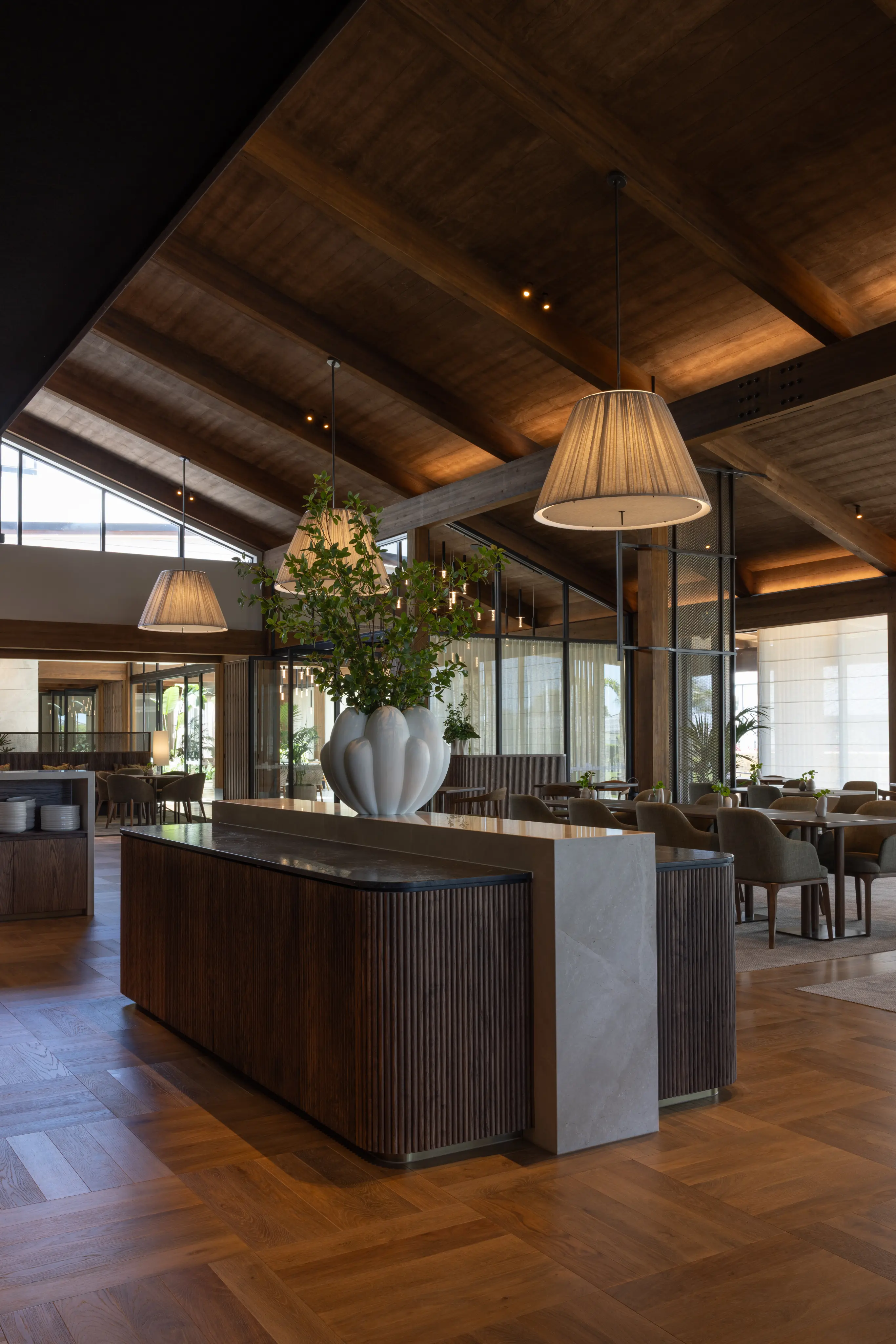 A large scalloped white vase of foliage on a stone-topped bar counter beneath pleated pendant lights in an open dining room.