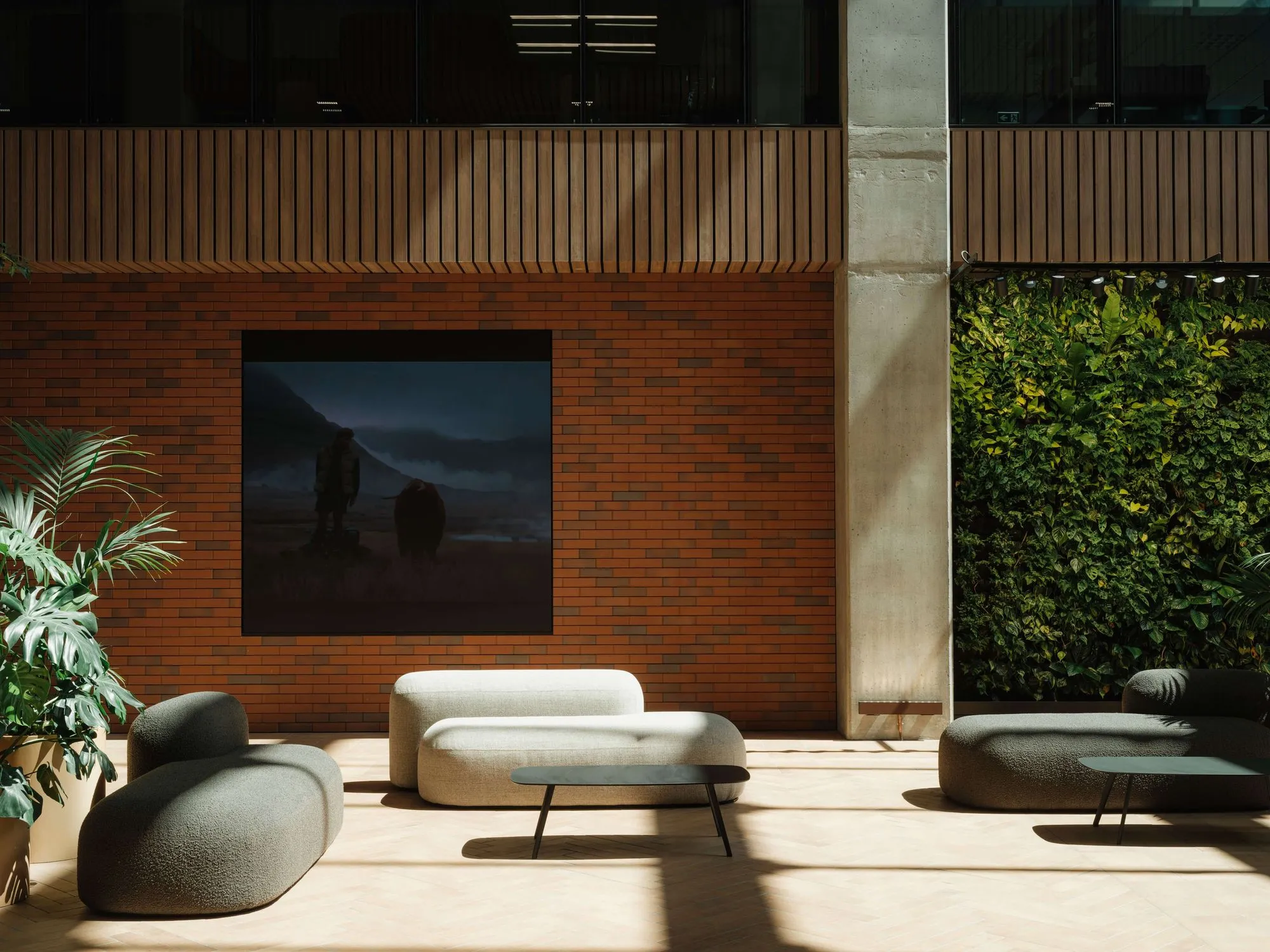 Kentia palm and Monstera beside sculptural lounge seating, with a green wall against red brick in the sunlit atrium.