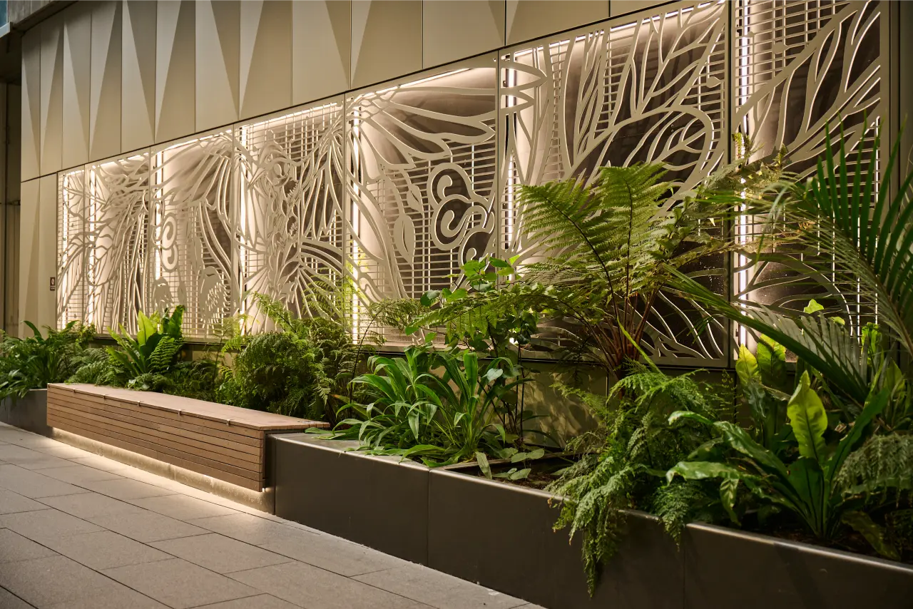 Layered fern planting along a timber bench beneath an illuminated bird artwork panel on the building facade.