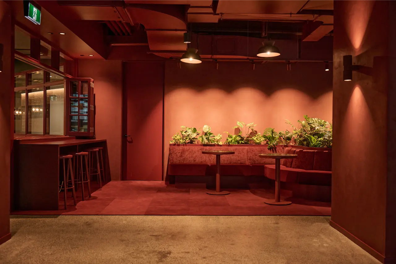 entertainment space wide, banquette planter, bar): Wide view of the Kernal Wealth entertainment space showing the custom inbuilt banquette planter of tropical foliage along the back wall, with bar seating and warm red-toned pendant lighting.