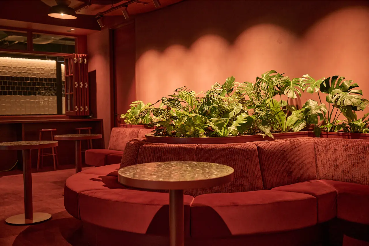 curved banquette and planter, wide entertainment space): Curved red velvet banquette seating with integrated planter of Monstera and Calathea in the Kernal Wealth entertainment space, lit by warm pendant and grow lighting.