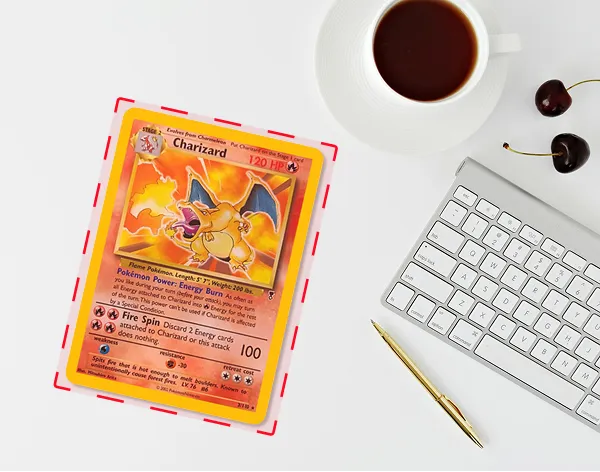 Charizard Pokémon card placed beside a white cup of coffee, two cherries, a gold pen, and a silver keyboard on a white surface.