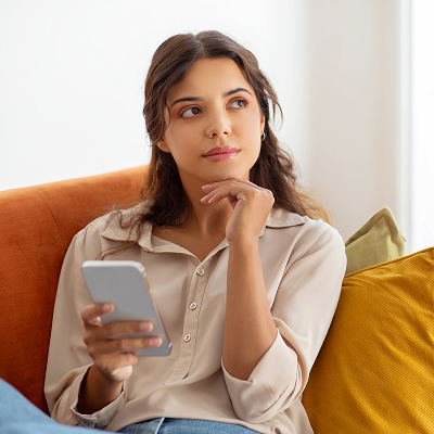 Pensive young woman holding smartphone and looking away while relaxing on couch at home