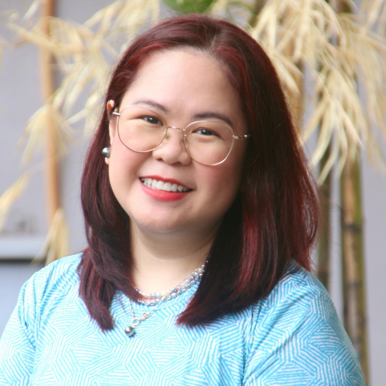 Smiling woman with shoulder-length dark red hair wearing glasses, red lipstick, pearl earrings, layered pearl necklaces, and a light blue patterned shirt, standing in front of blurred yellow-green plants.