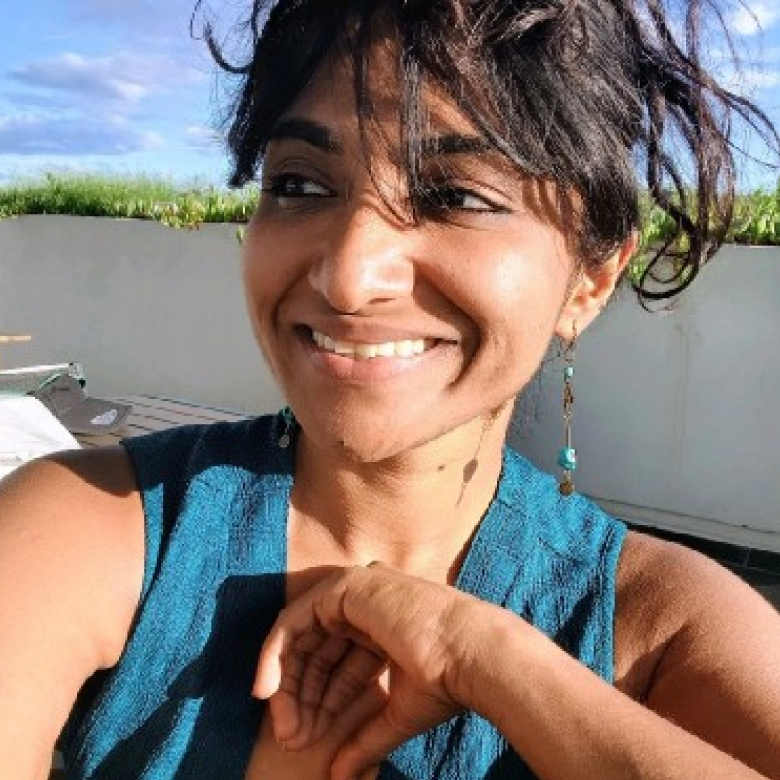 Smiling woman with dark hair loosely tied, wearing blue sleeveless top and dangling turquoise earrings, outdoors with blue sky and greenery in the background.